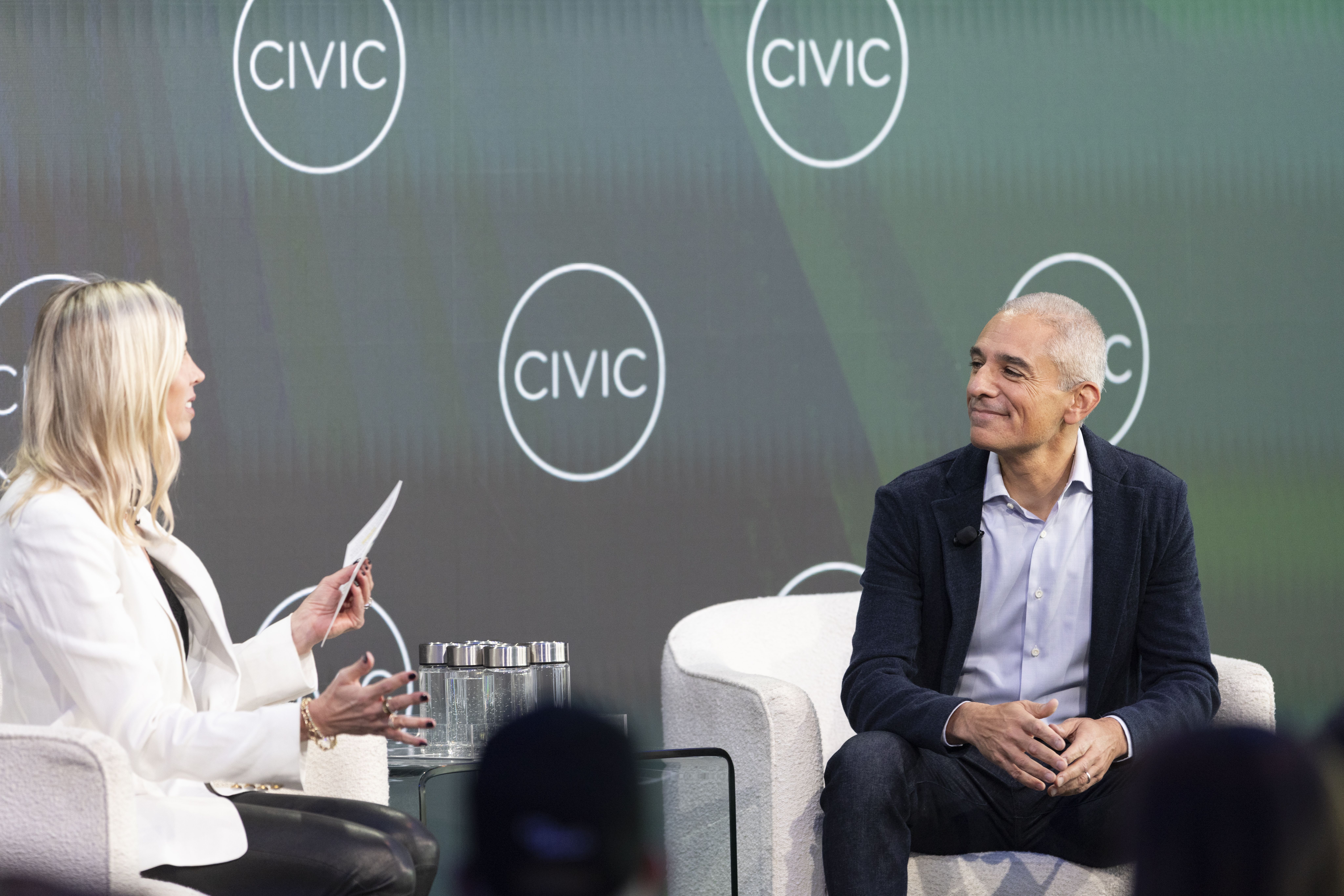 Two people in discussion on a stage. Left: a woman in a white blazer; right: a man in a dark blazer. Green backdrop with white circular CIVIC logos and a glass table with water.