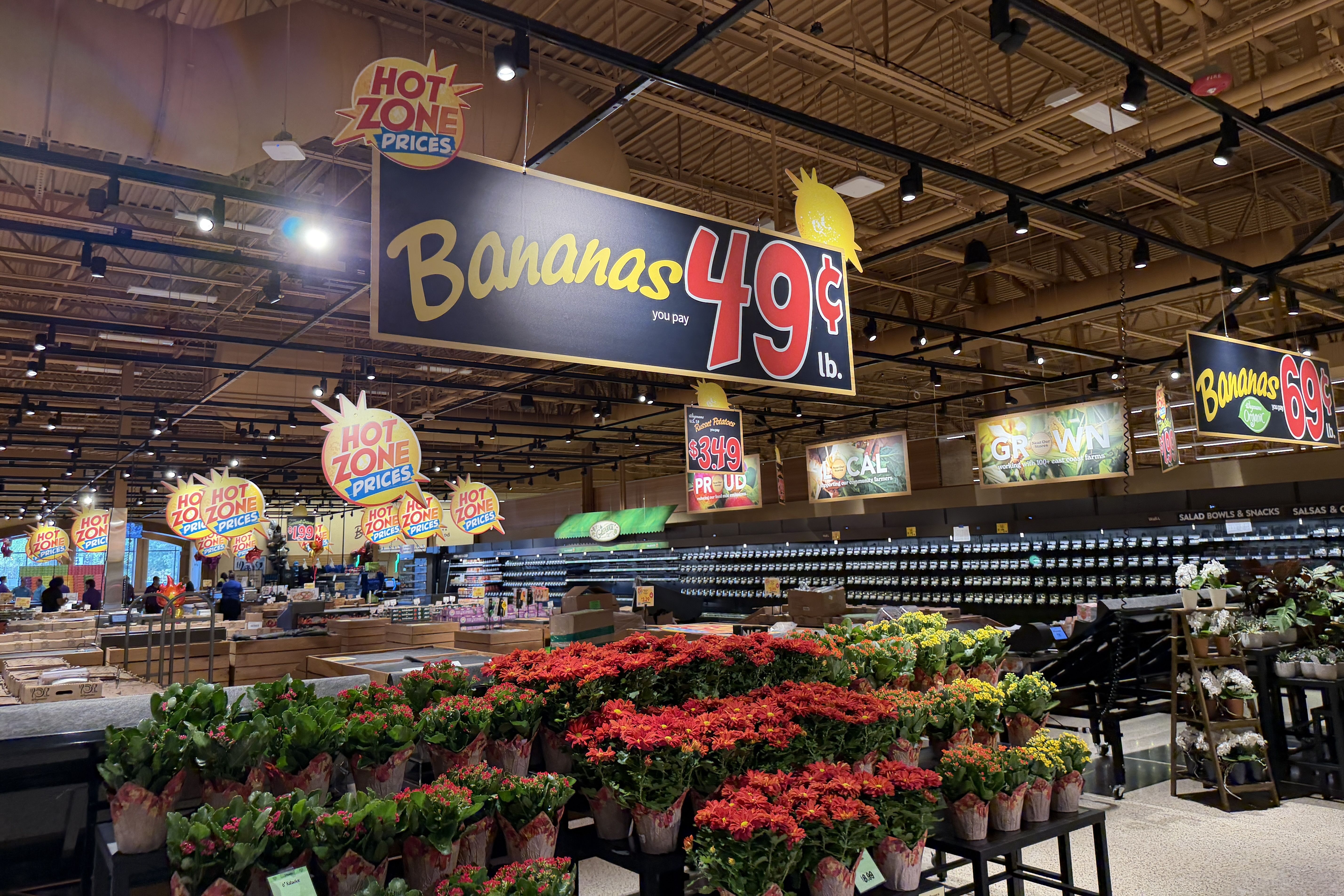 Grocery store interior with a large blue sign reading "Bananas 49¢/lb" and a pineapple graphic; overhead signs say "Hot Zone Prices" as rows of colorful potted flowers line the foreground.