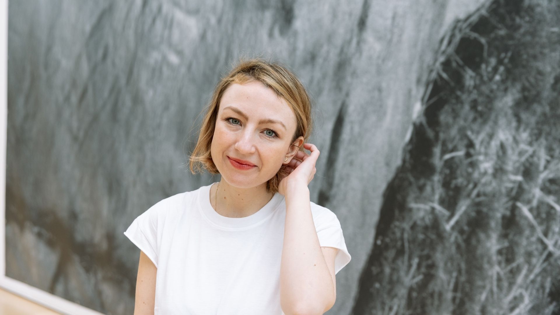 Portrait of a woman with short blond hair wearing a white shirt, smiling softly and touching her hair, standing before a gray marble wall.
