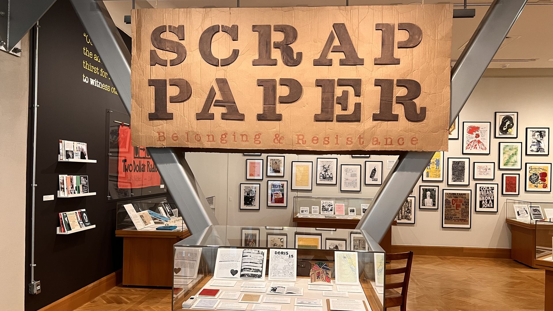 Large crumpled brown banner reading "SCRAP PAPER" in bold letters dominates the display, with "Belonging & Resistance" faint beneath; walls feature framed prints and a glass display case.