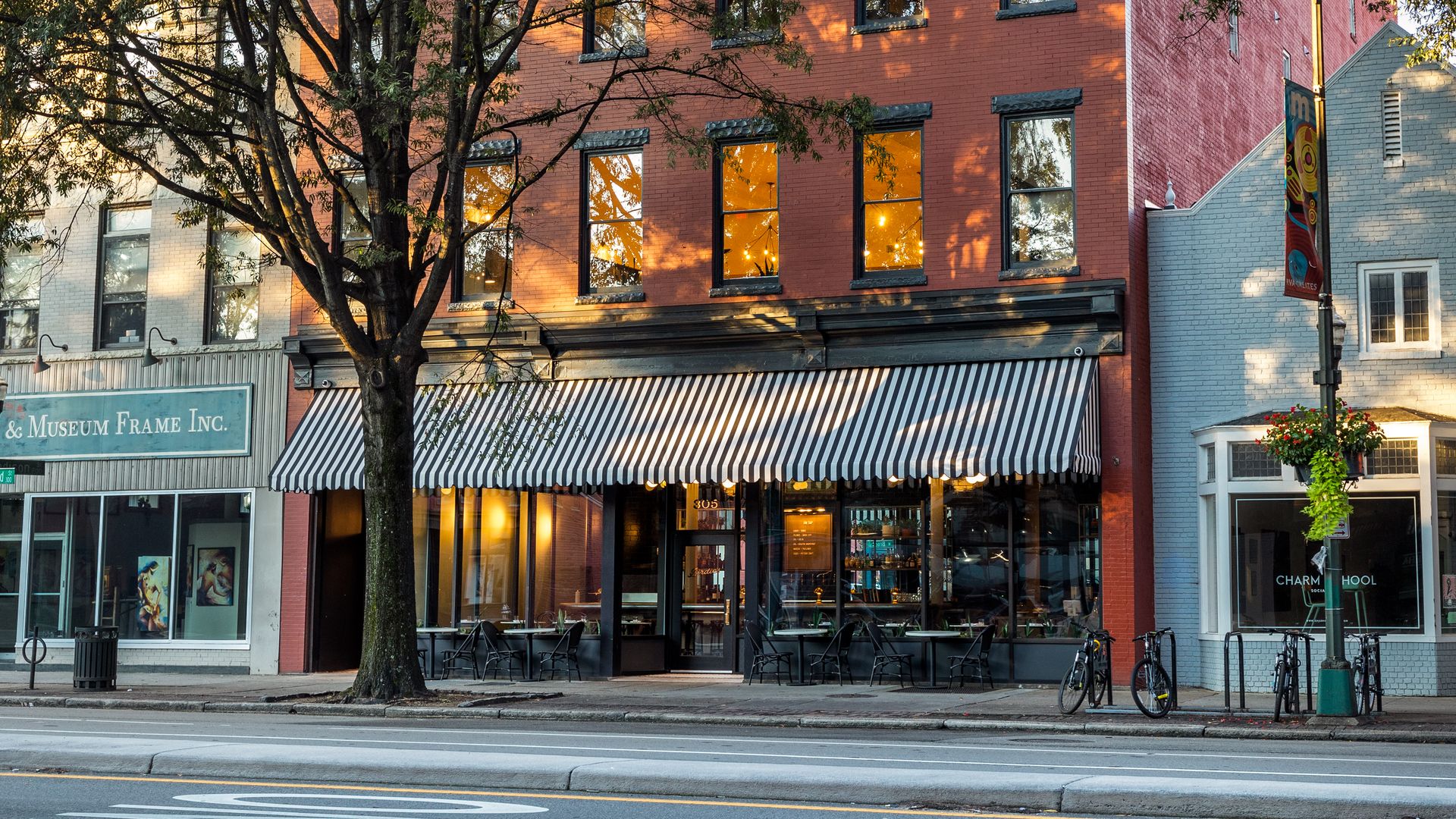 Red brick urban building with lush trees in front; ground-floor shop under a black-and-white striped awning, warm-lit windows, and bicycles parked along the curb on a sunny day.