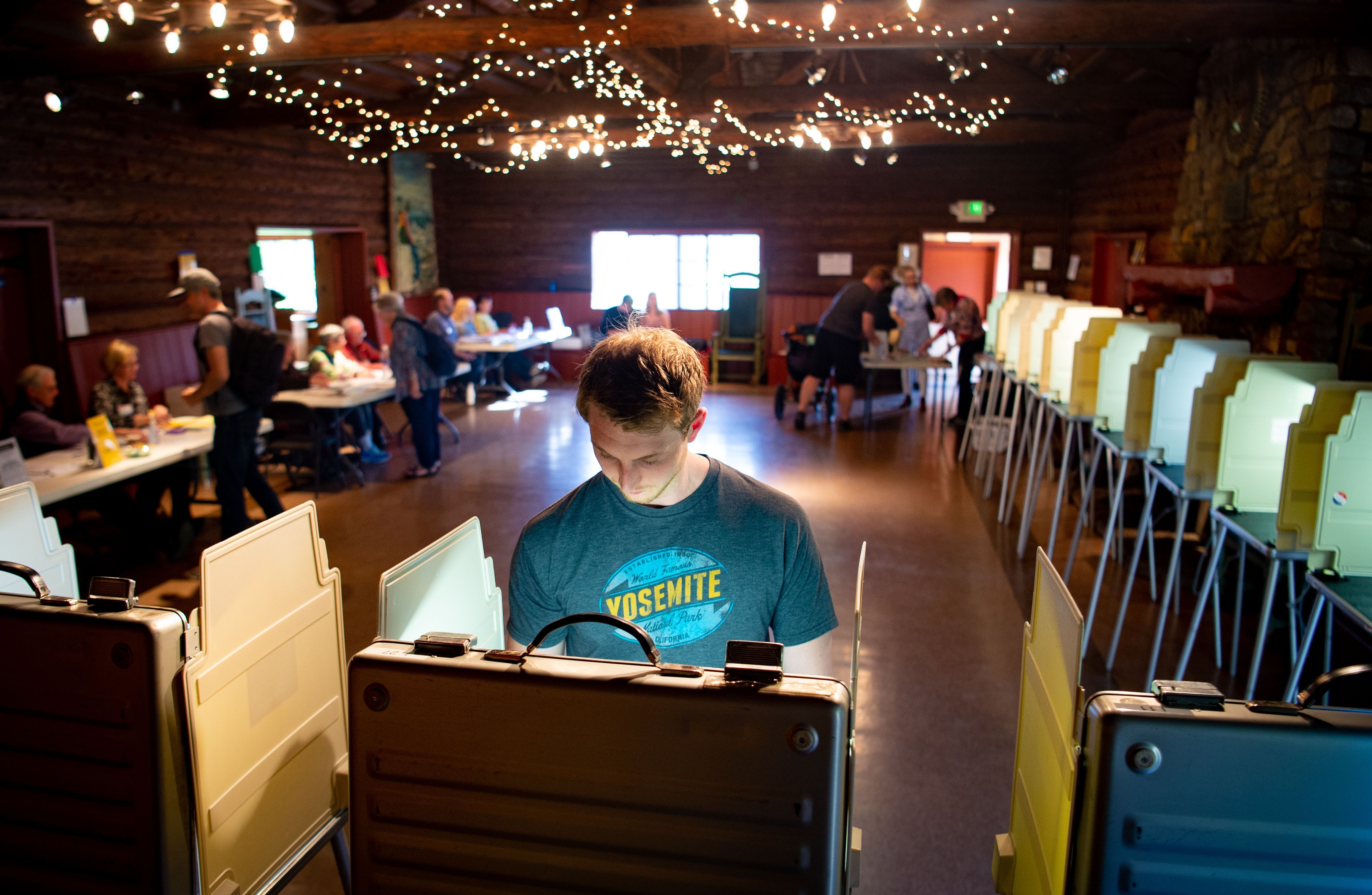 In this image, a man stands at a voting booth in. room with fairy lights on the ceiling 