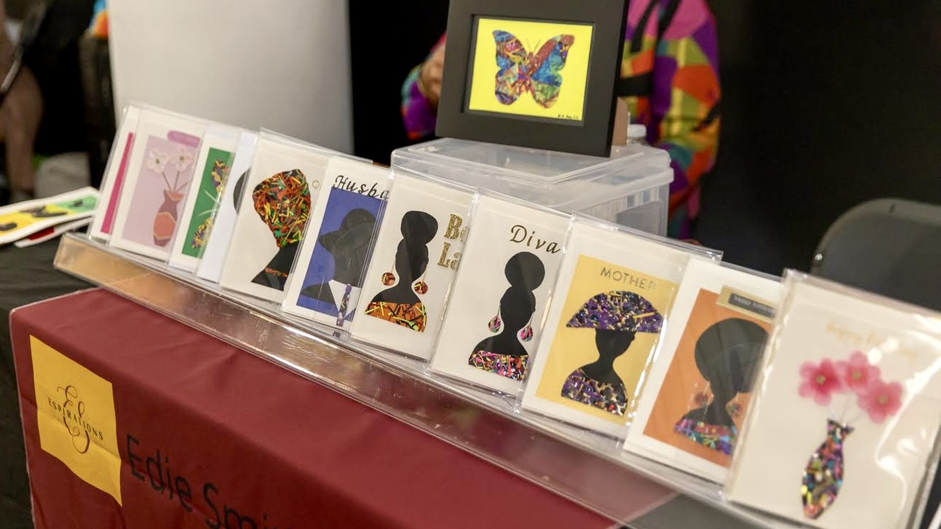 Display of colorful greeting cards featuring black female silhouettes, floral designs, and a butterfly artwork on a table with a burgundy cloth labeled Edie Smith Inspirations.