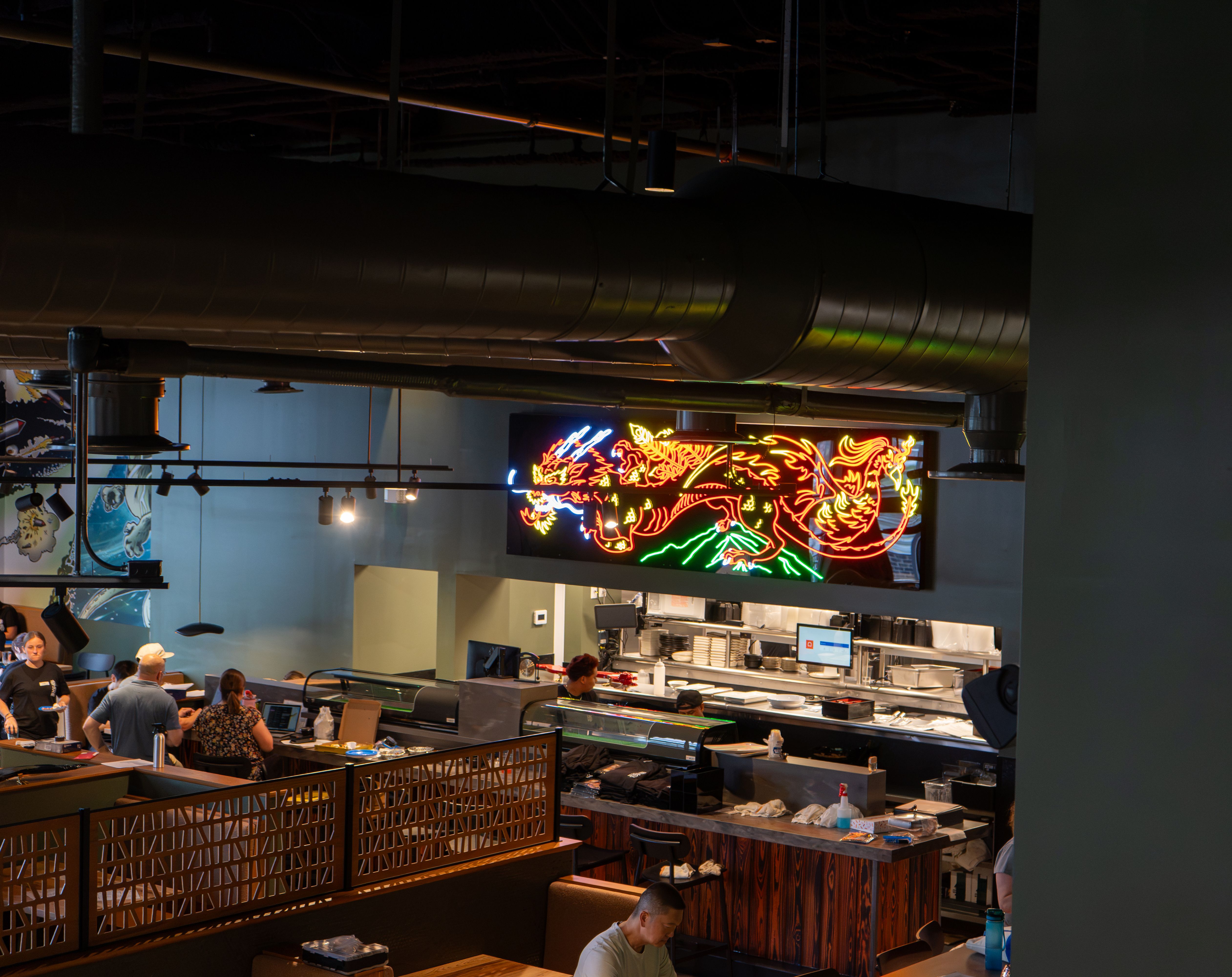 Dim restaurant interior with exposed ducts and pipes on the ceiling; a bright neon dragon sign glows over the counter as patrons sit at tables and the front counter area.