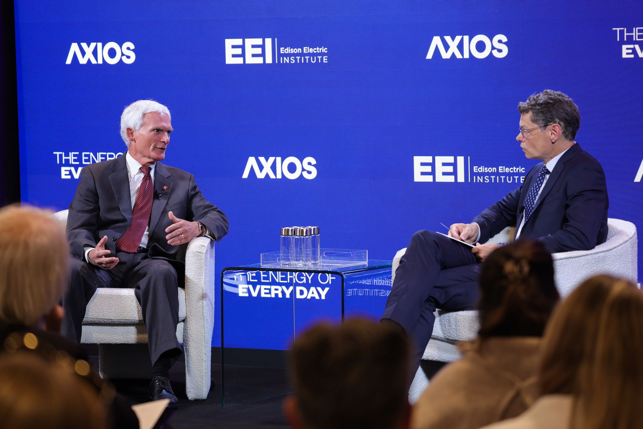 Two men in suits on a stage with a blue backdrop showing AXIOS and Edison Electric Institute logos. They speak on a panel; a glass table with water glasses sits between them; audience in foreground.