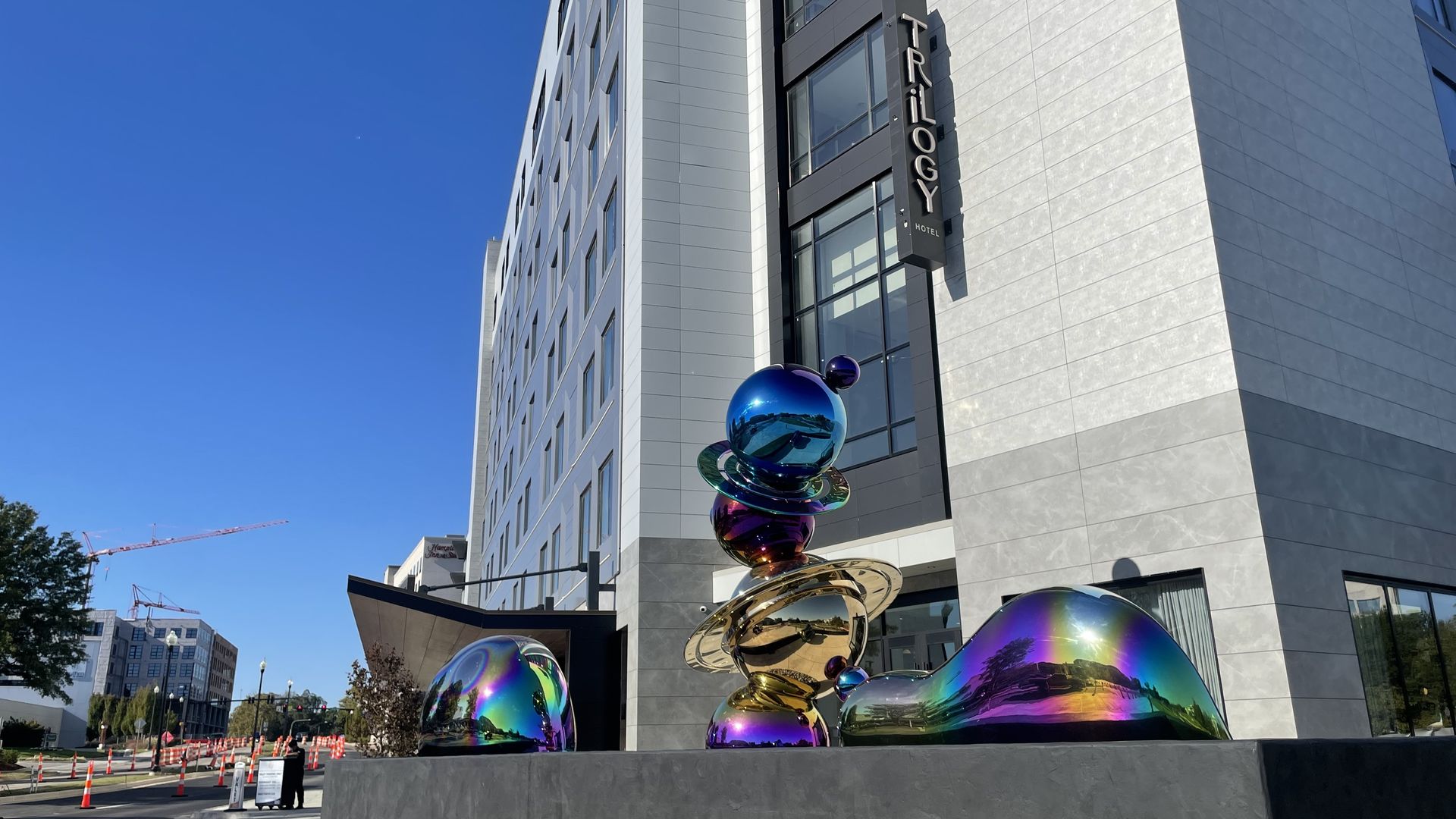 Iridescent, reflective abstract sculptures in front of the modern white and gray Trilogy Hotel building under a clear blue sky.