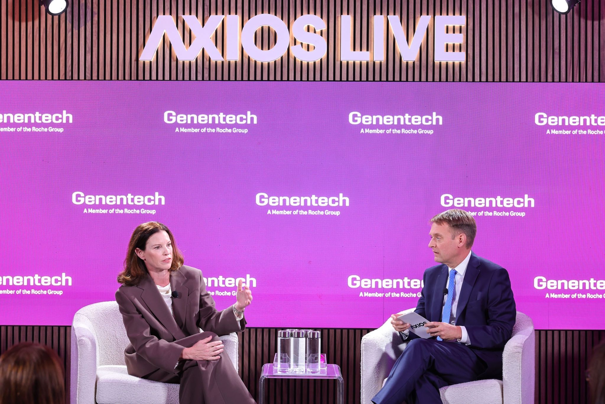 Two speakers in suits sit on white chairs on a stage with a purple Genentech backdrop and a large "AXIOS LIVE" sign overhead; the woman speaks while the man listens.