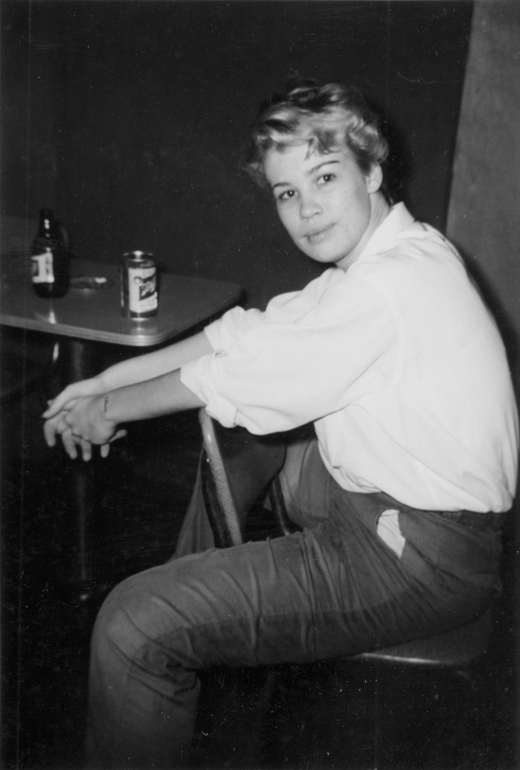 A black and white photograph of a light-haired person sitting backwards on a chair at Jimmie White's Tavern; they are smiling softly at the camera. 