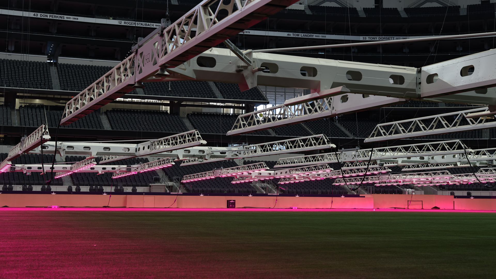 Inside a large empty sports arena, white steel trusses sweep overhead while pink lights glow on the floor; dark seating surrounds, and banners with names and years line the upper walls.