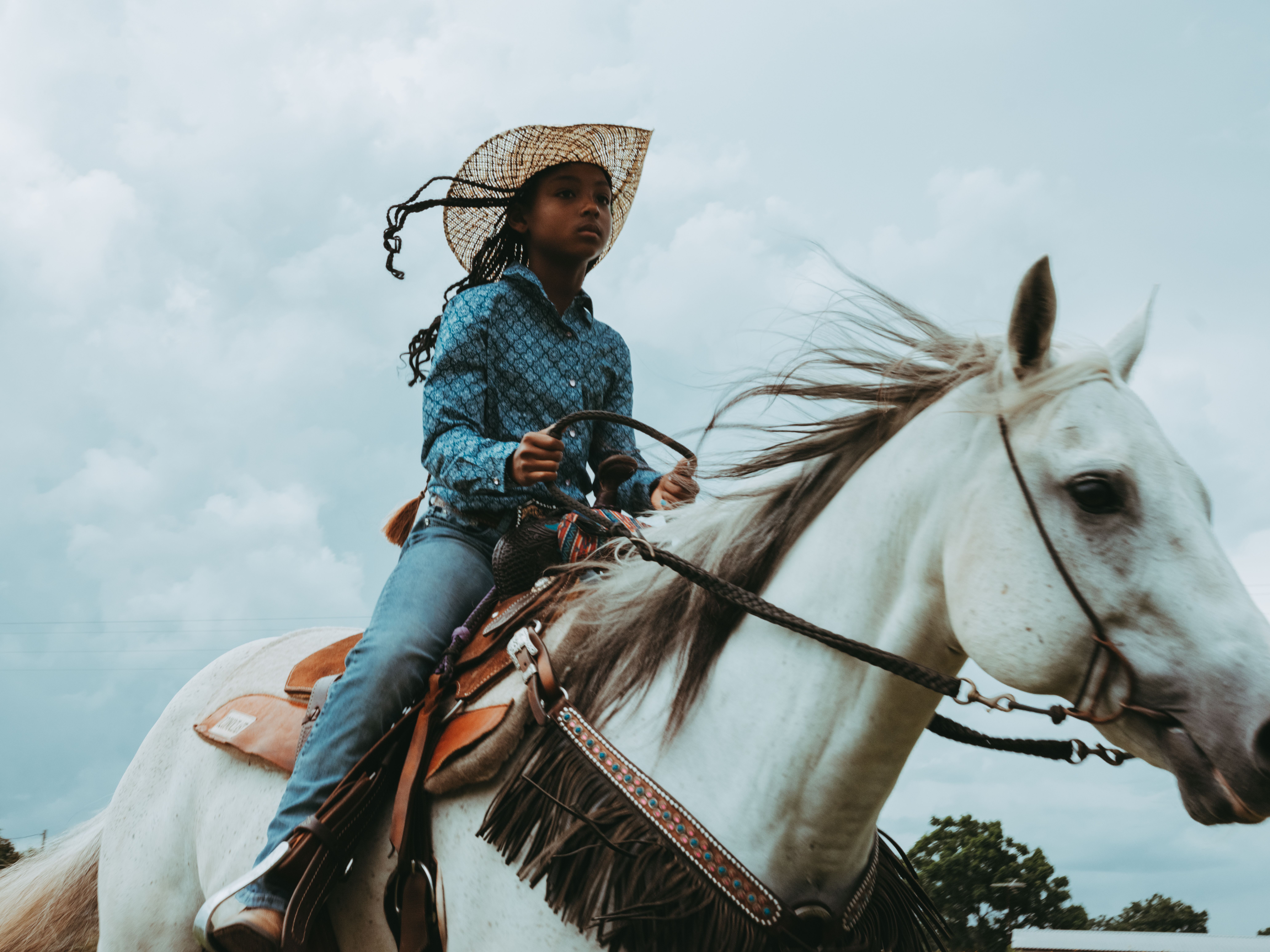 A Black cowgirl with braids rides a white horse.