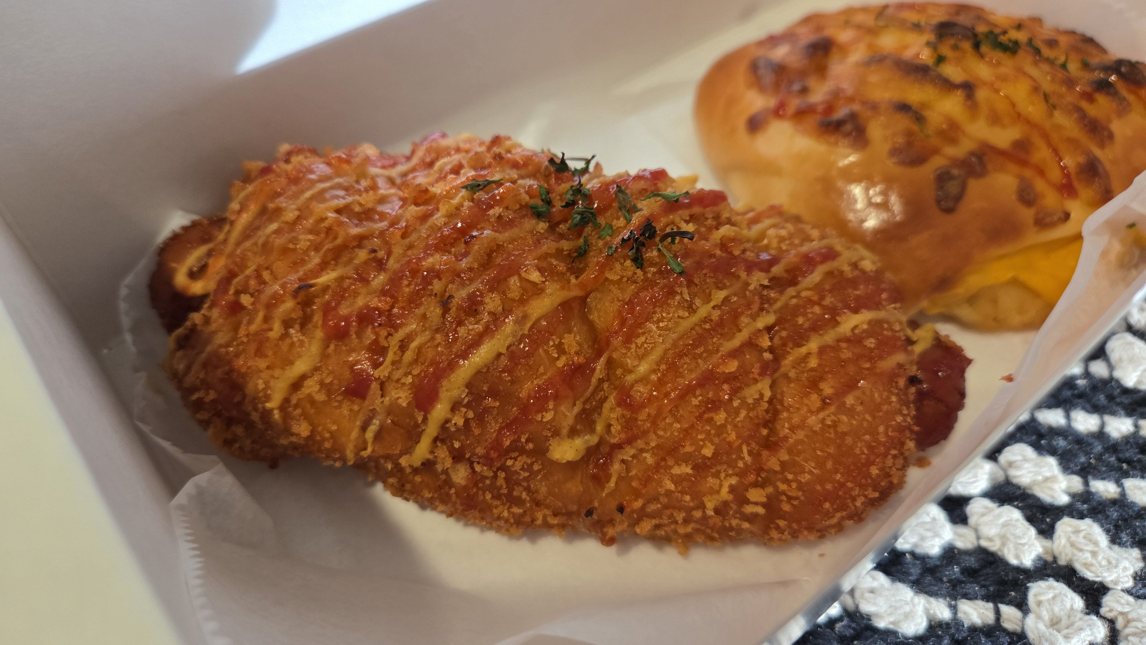 Golden breadcrumb-coated fried food in a white takeout box; the front piece is glazed with a reddish sauce and sprinkled with chopped parsley, with a cheesy baked roll visible in the background.