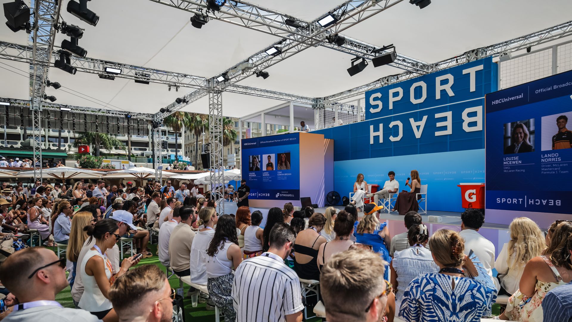 Outdoor event with a large audience, blue stage backdrop reading "SPORT BEACH", metal truss lighting above and speakers at microphones; sunny day with umbrellas and palm trees.