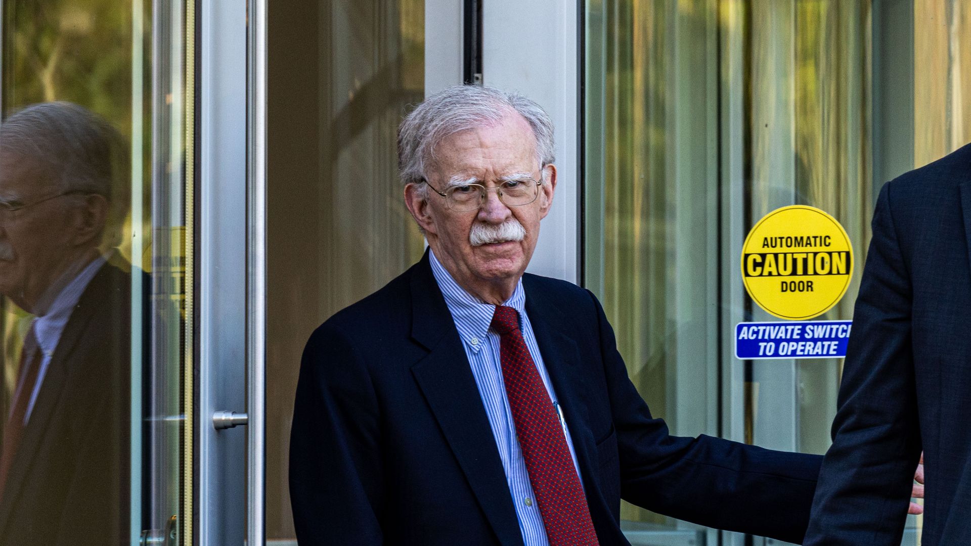 John Bolton leaves the federal court building wearing a navy jacket, blue shirt and red tie. 