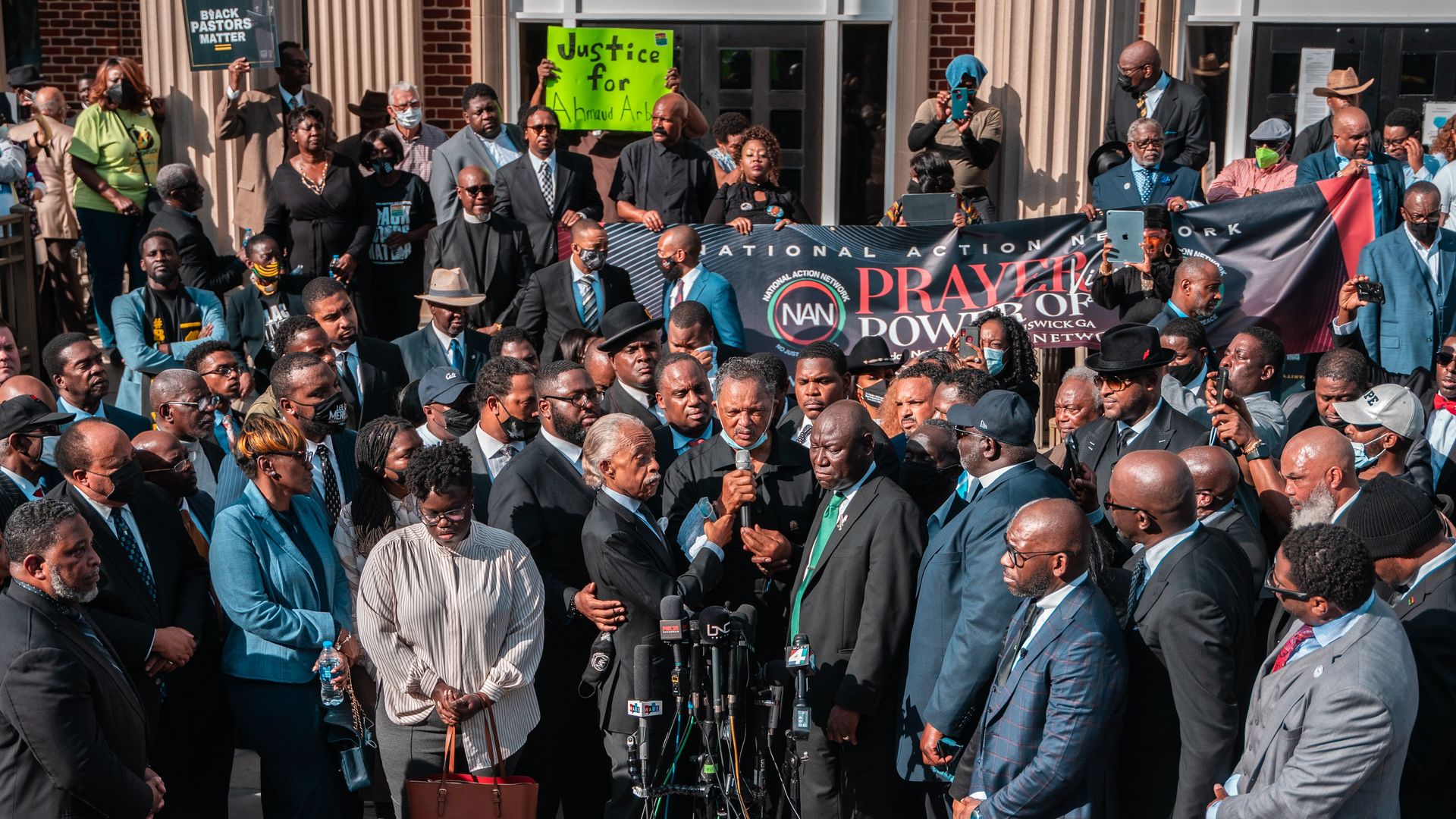 A man speaks into a microphone, surrounded by dozens of others in front of a courthouse