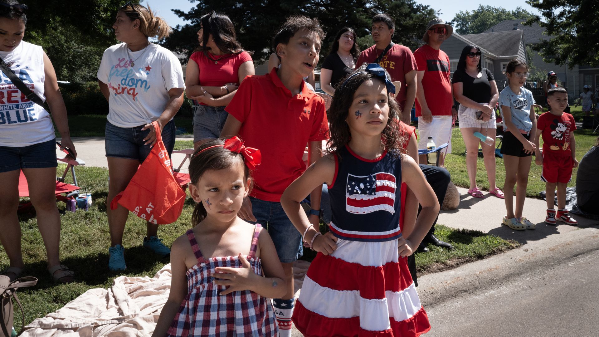 Three children with different expressions wearing red, white and blue stand on the side of a street with other onlookers behind them.