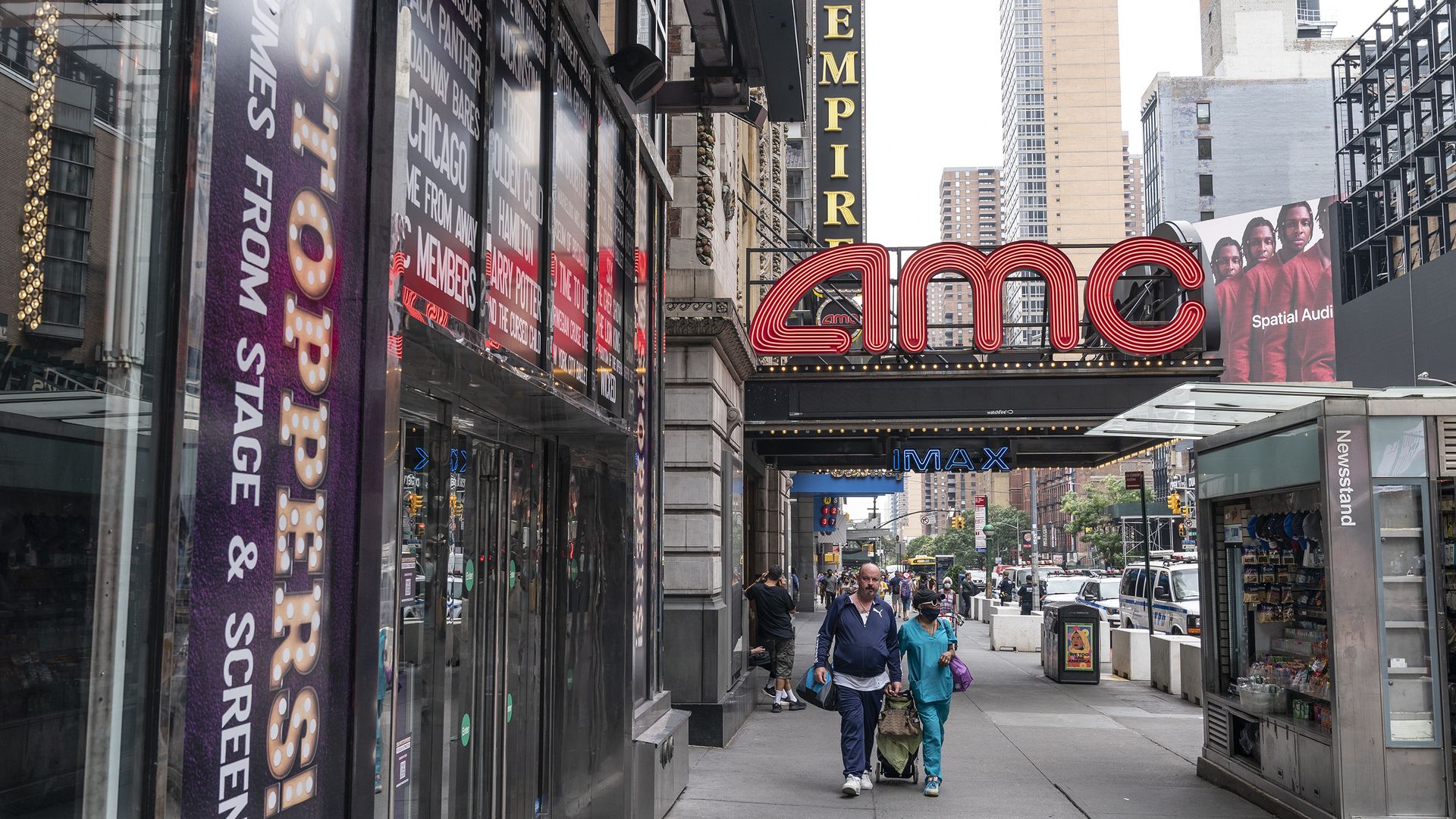 An AMC sign hangs above walkers along a sidewalk in an urban environment.