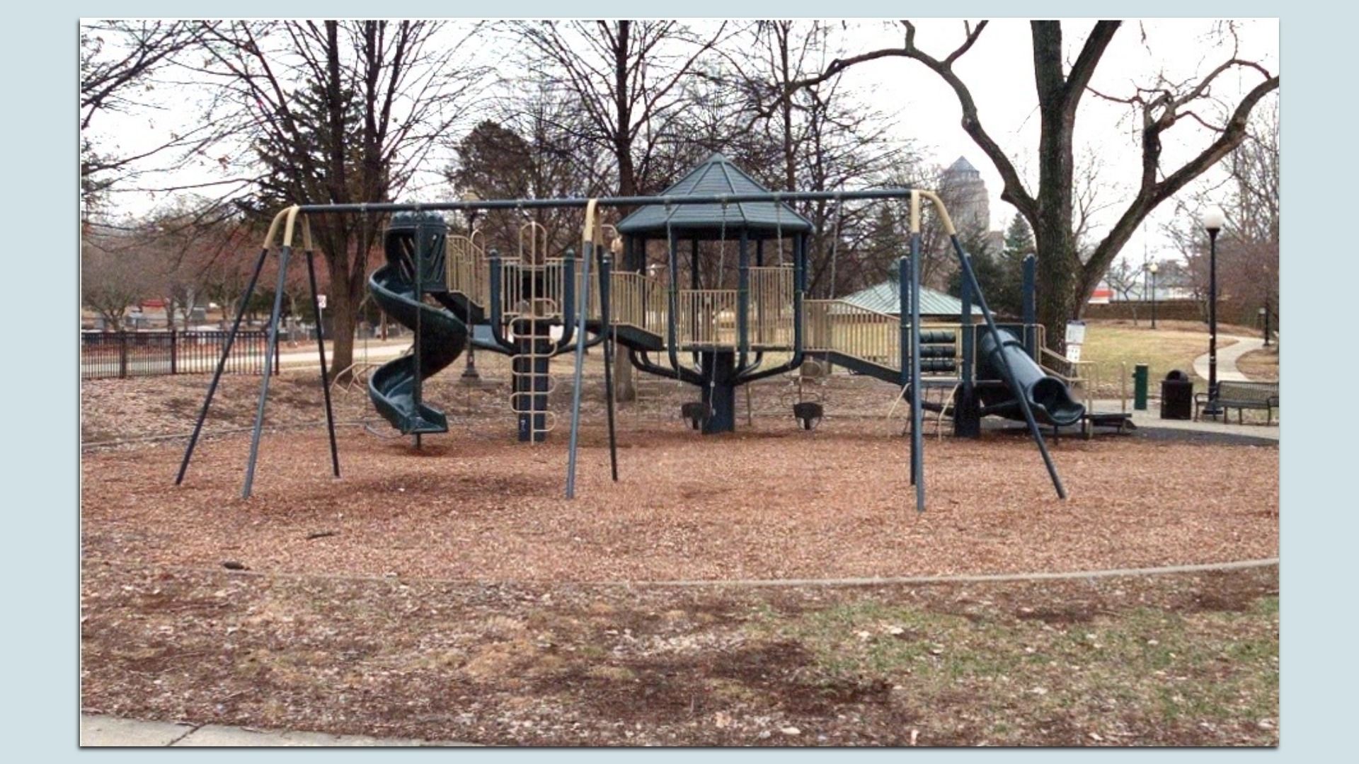 A playground with blue and tan equipment, including a spiral slide, straight slides, ladders, and a central covered tower, set on wood-chip mulch among bare trees in a park.