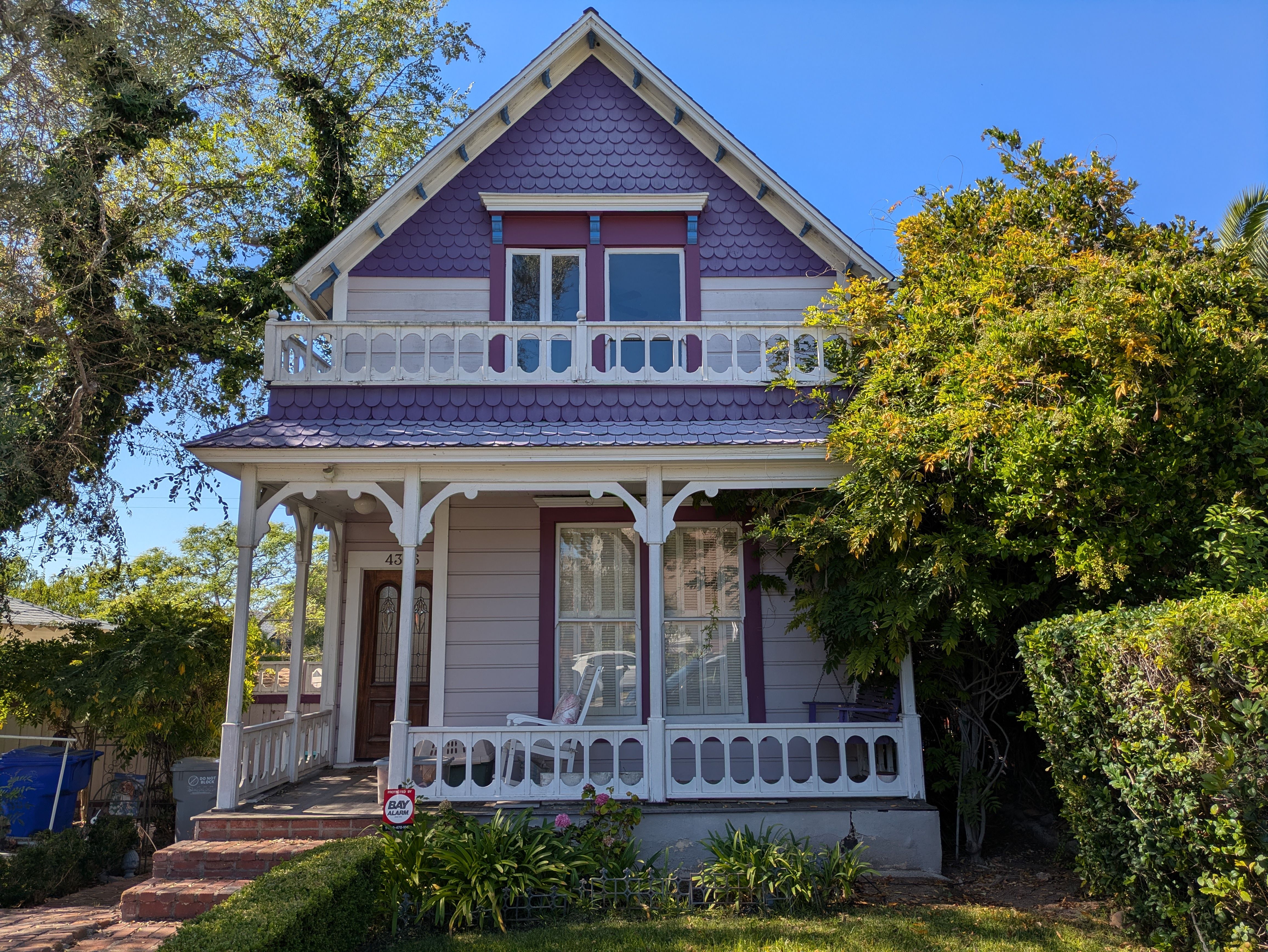 Two-story house with purple scalloped siding, white trim, a covered porch, and greenery on the right under clear blue sky.