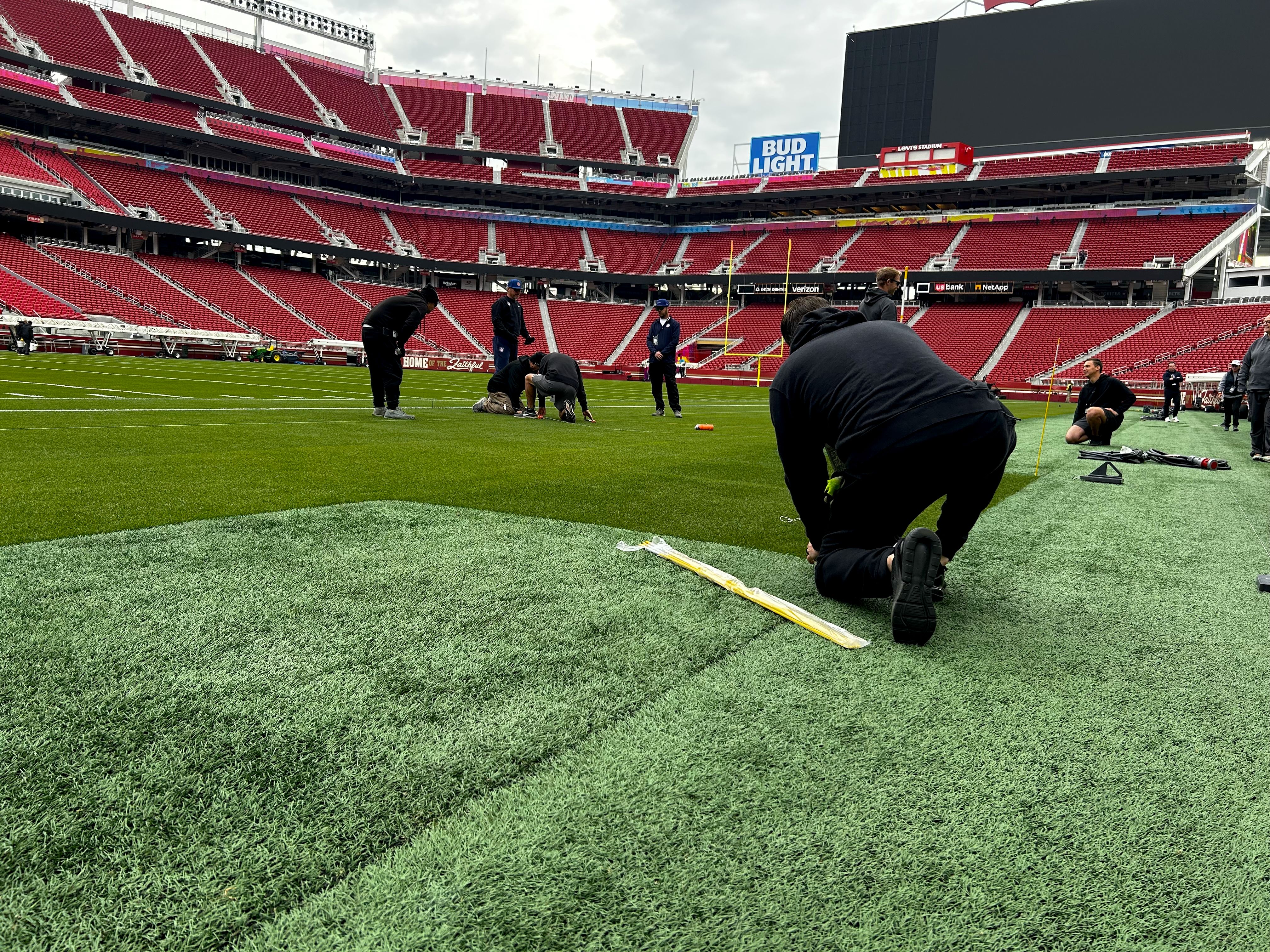Workers kneeling and standing on a green football field inside a large stadium with red seats, installing or repairing turf near the sidelines on a cloudy day.