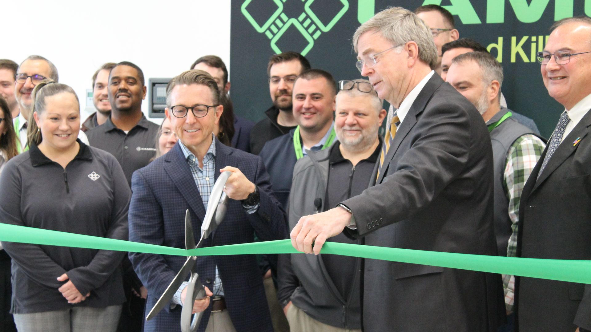 Group of people smiling at a ribbon-cutting event. A man in a blue blazer cuts a green ribbon with oversized scissors, helped by a man in a black suit and striped tie, with others around them.