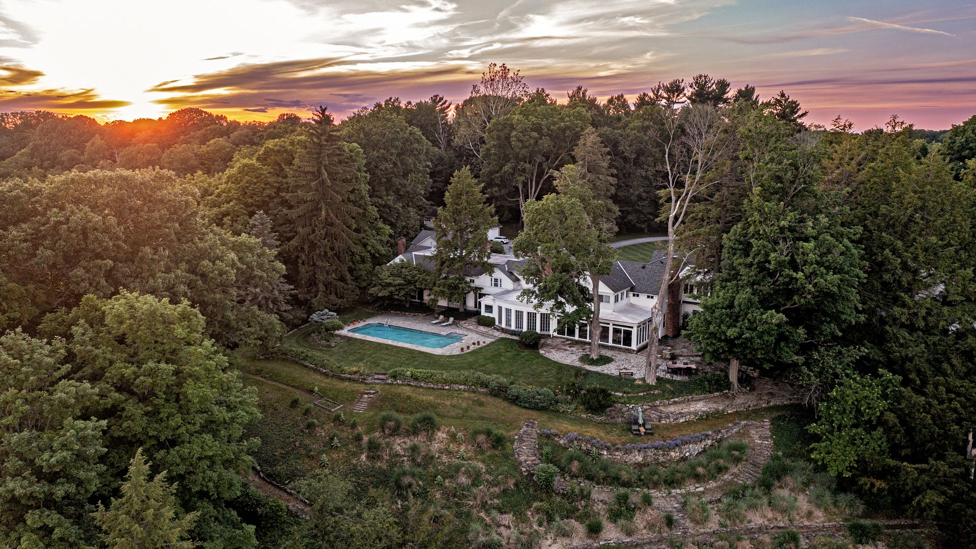 Aerial view of a white house with a rectangular pool in a wooded backyard, surrounded by tall trees as a sunset blooms in orange, pink, and purple across the sky.