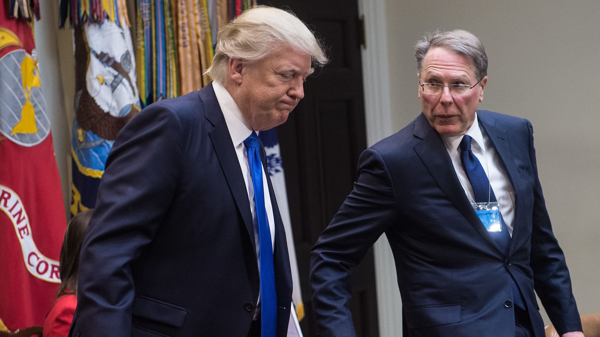 In this image, Trump and LaPierre pull out chairs to sit next to each other at a table. Both are wearing suits.