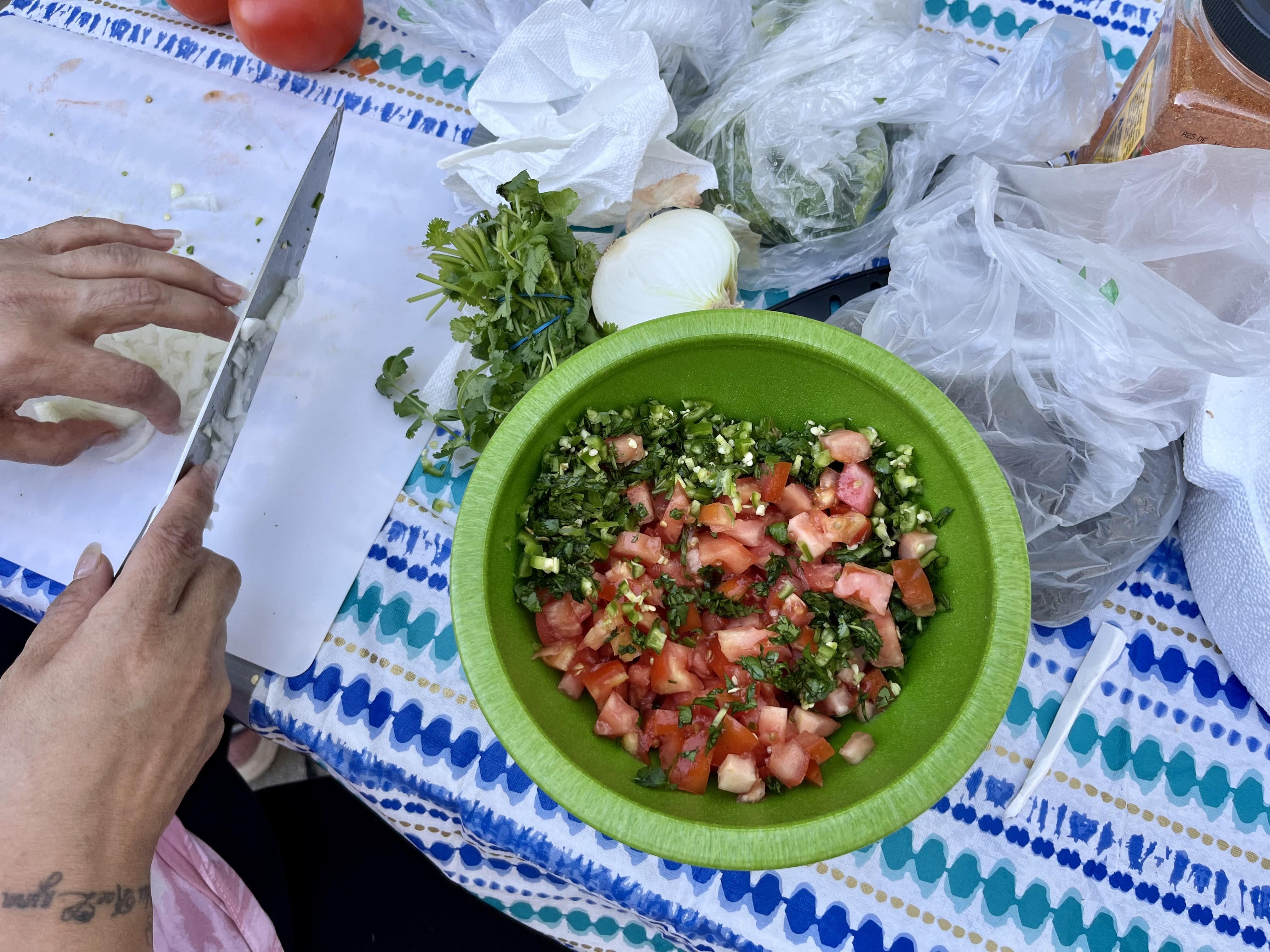 A bright green bowl on a blue tablecloth holds bright red tomatoes and green peppers. A woman uses a knife to dice onion in the corner.