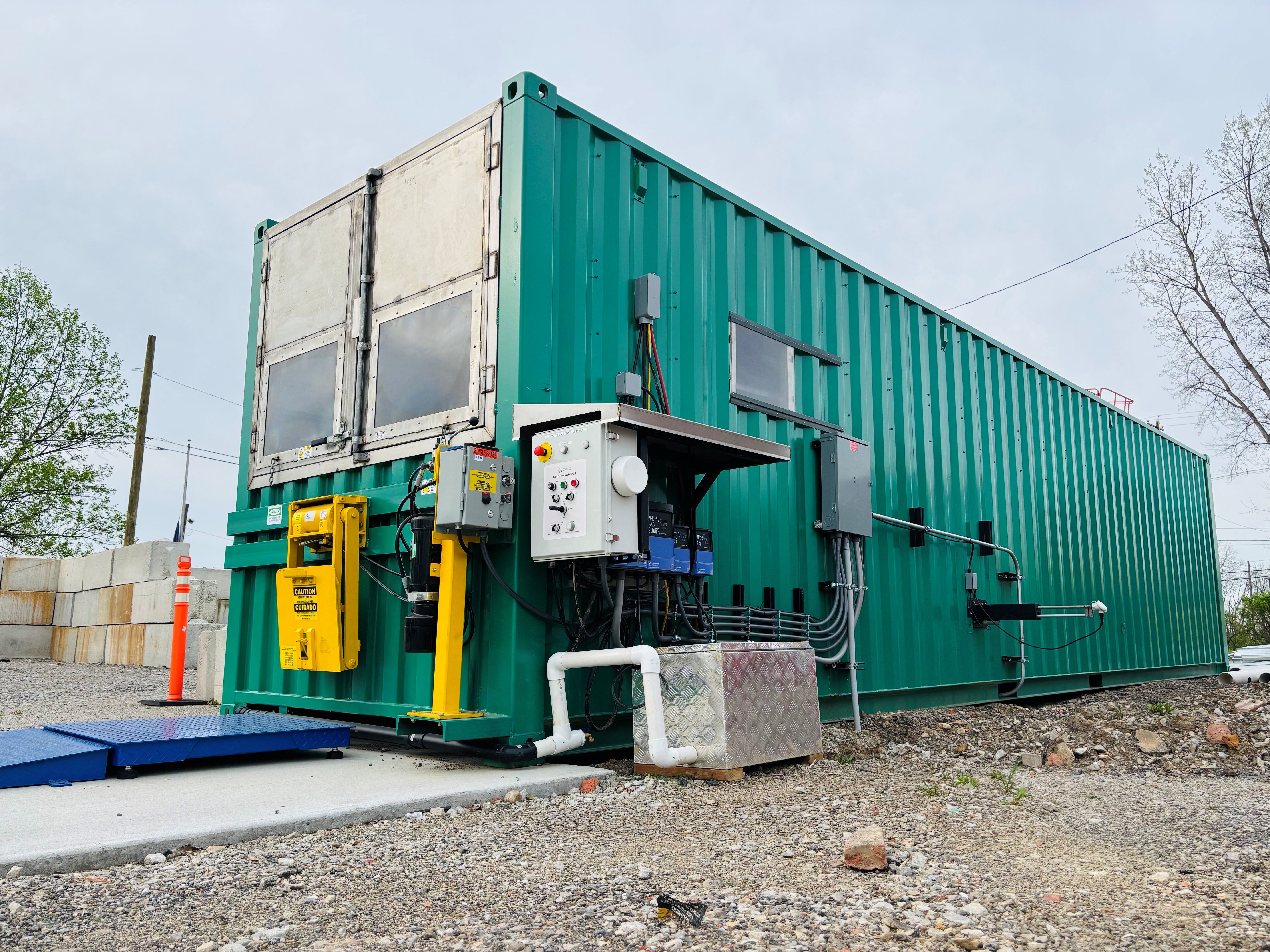 Green modular container with large gray doors and exposed electrical panels; colorful wires run along its side. Yellow safety system, blue ramp, gravel ground, leafless trees and pale sky.
