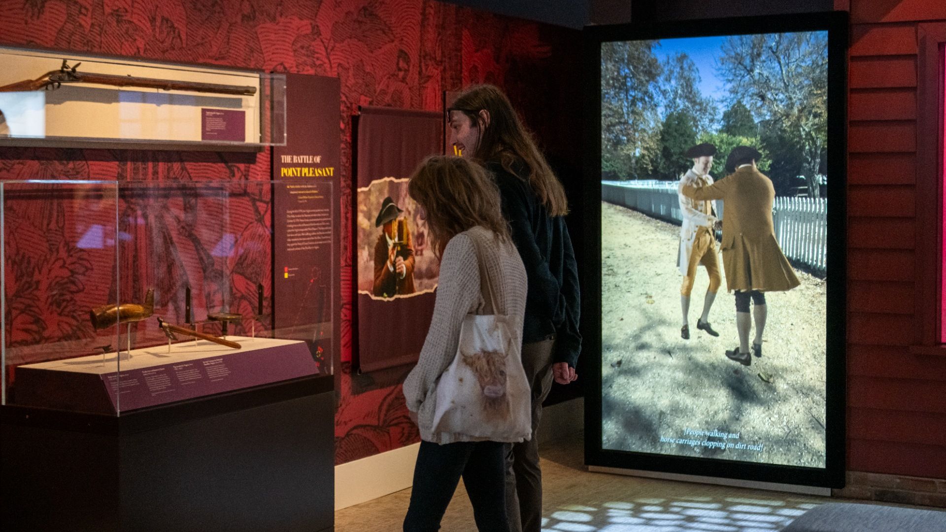 Two people view a museum exhibit about the Battle of Point Pleasant with rifles in glass cases and a digital display showing two men in 18th-century attire outdoors.