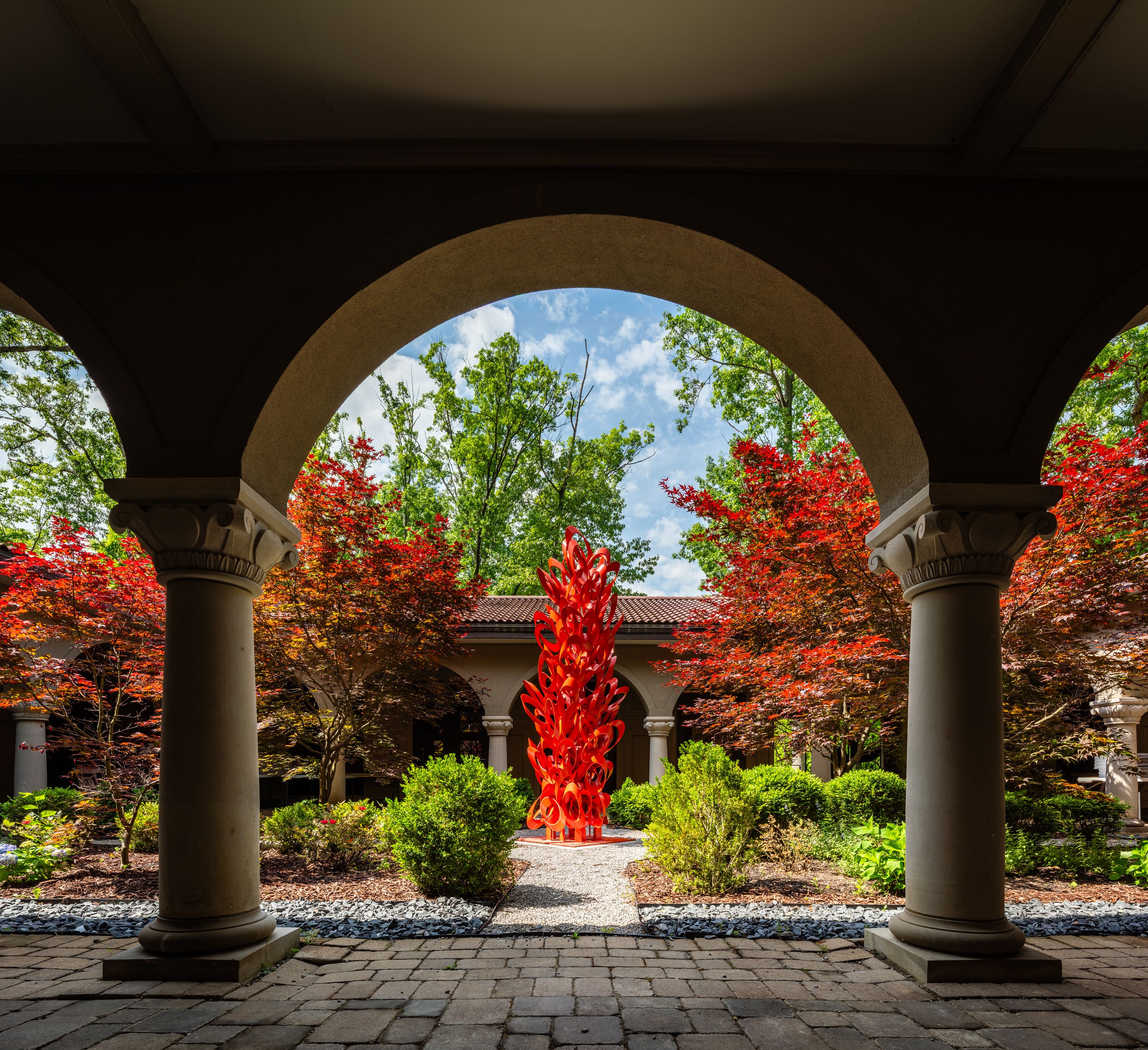 Under a dark arched walkway, beige columns frame a tall bright red glass sculpture in a sunlit courtyard, with red and green foliage, blue sky, and a stone-paved path.