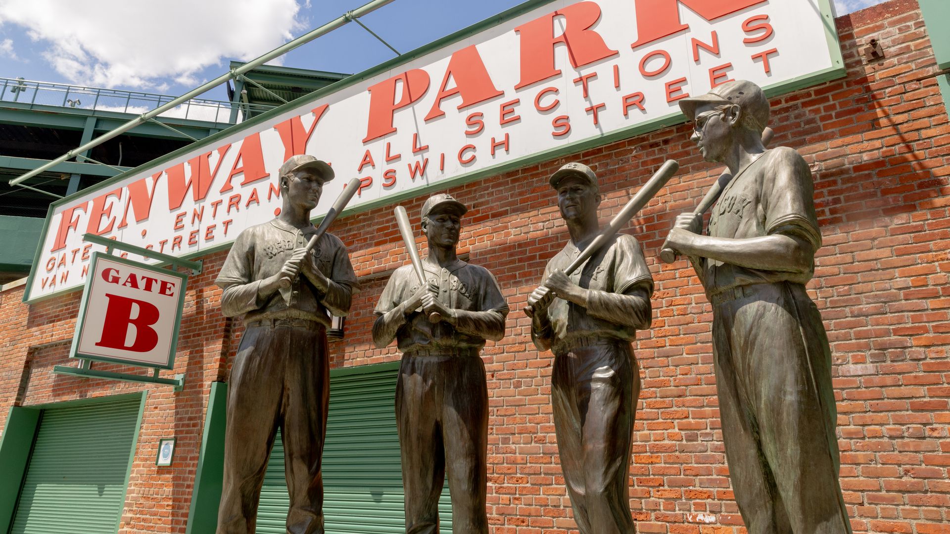 Bronze statues of four baseball players holding bats outside Fenway Park near Gate B, red brick wall and clear blue sky with white clouds in the background.