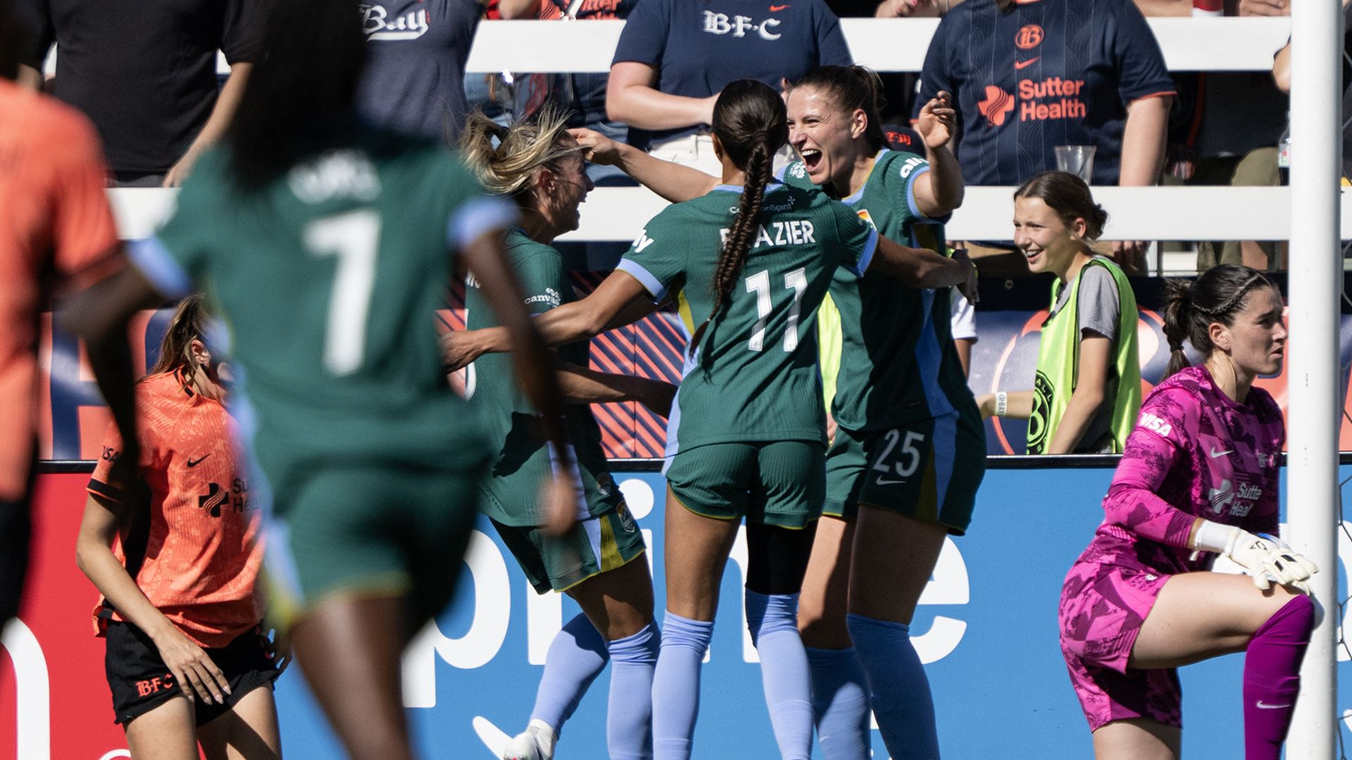 Soccer players in teal uniforms celebrate a goal near the net as teammates embrace; a pink-clad goalkeeper sits by the post, with cheering fans in the stands.