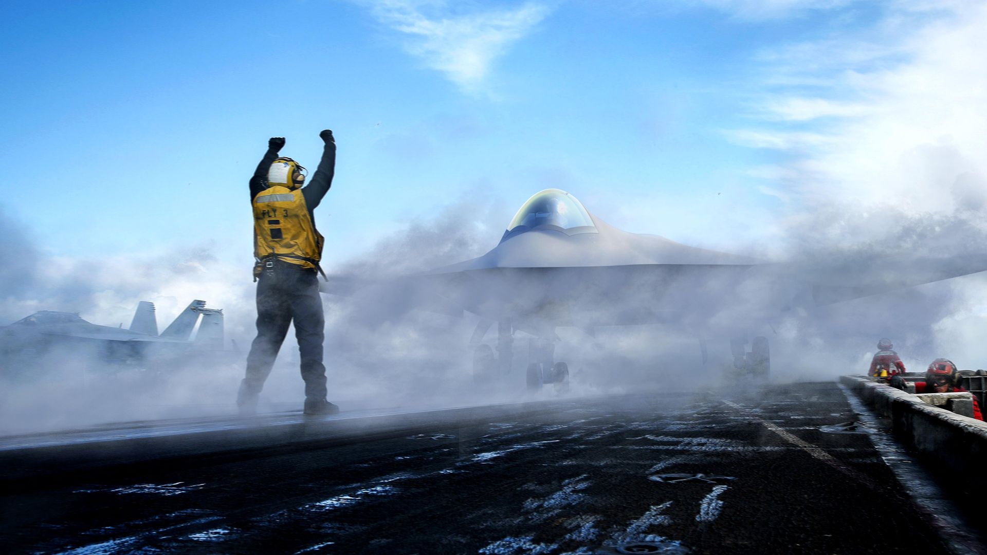A person in a yellow vest and wearing a helmet does hand signals toward a futuristic looking plane cloaked in smoke.