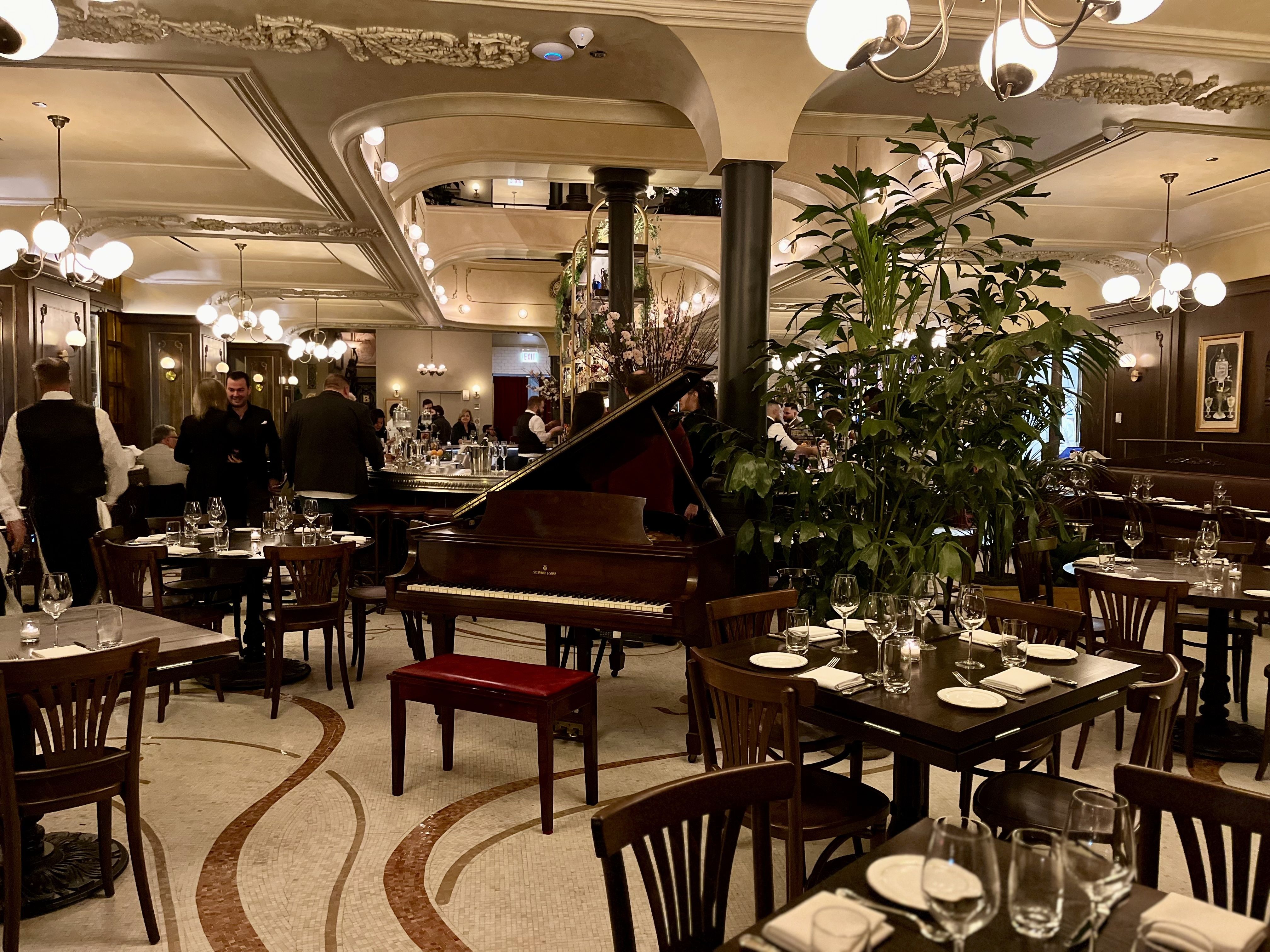 Elegant restaurant with a grand piano center stage, wooden tables set with glasses and plates, hanging globe lights, and tall green plants. Guests dine as staff mingle in the warmly lit space.