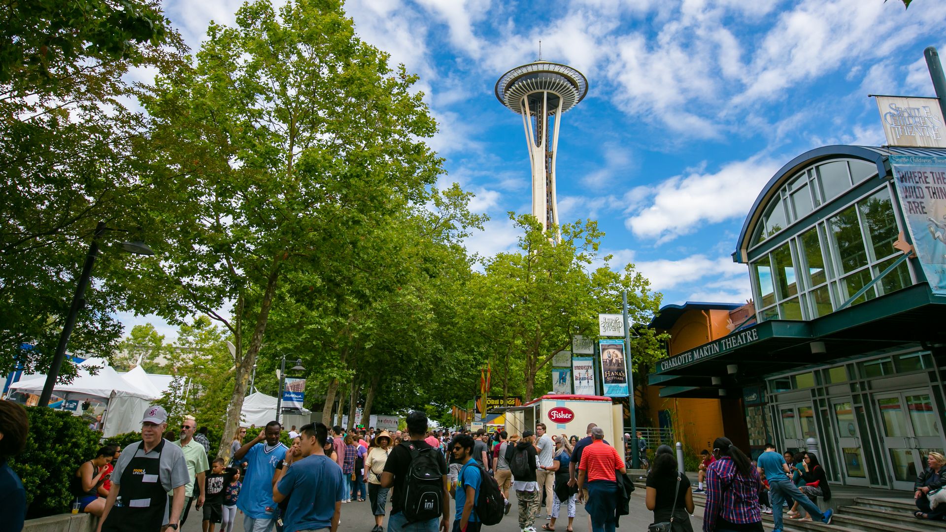 The Space Needle is shown over a crowd of people at Seattle Center, with blue skies punctuated by only a few clouds.