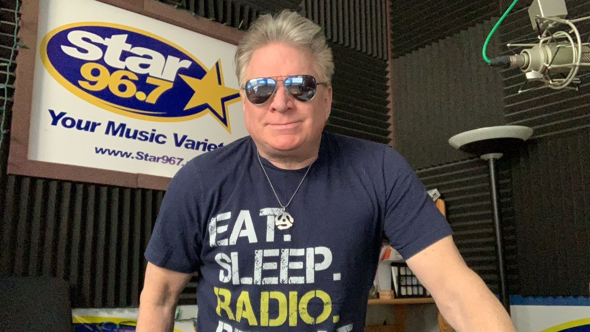 Smiling man wearing aviator sunglasses and a navy T-shirt that reads EAT. SLEEP. RADIO. REPEAT. stands in a radio studio with a Star 96.7 sign, a microphone, and acoustic foam panels behind him.