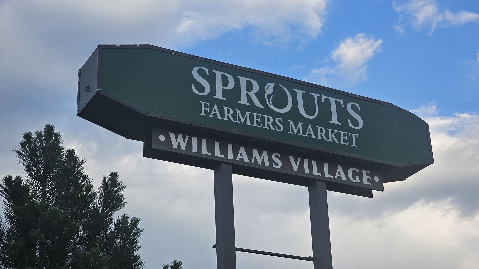 Green Sprouts Farmers Market sign on posts with large white lettering 'SPROUTS' and 'FARMERS MARKET'; 'WILLIAMS VILLAGE' partially visible, blue sky with clouds, foreground pine trees.