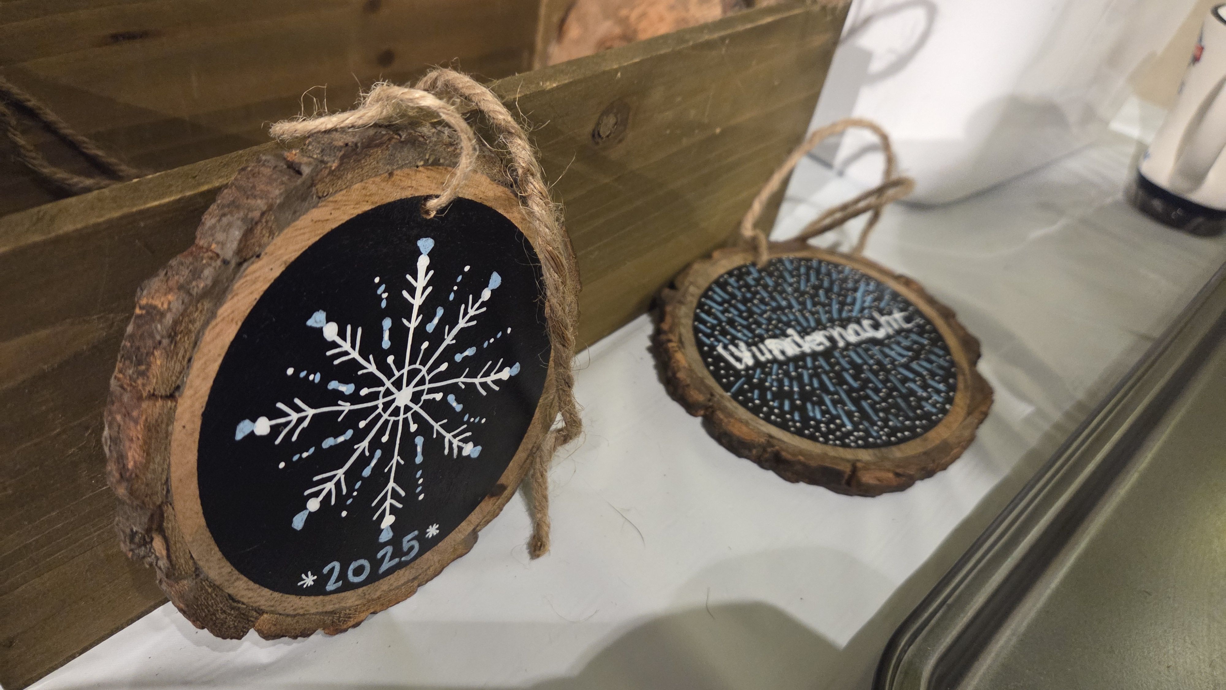 Two round wooden ornaments with bark edges, one painted with a white and blue snowflake and "2025", the other with blue strokes and the word "Winternacht" in white, both with hanging ropes.
