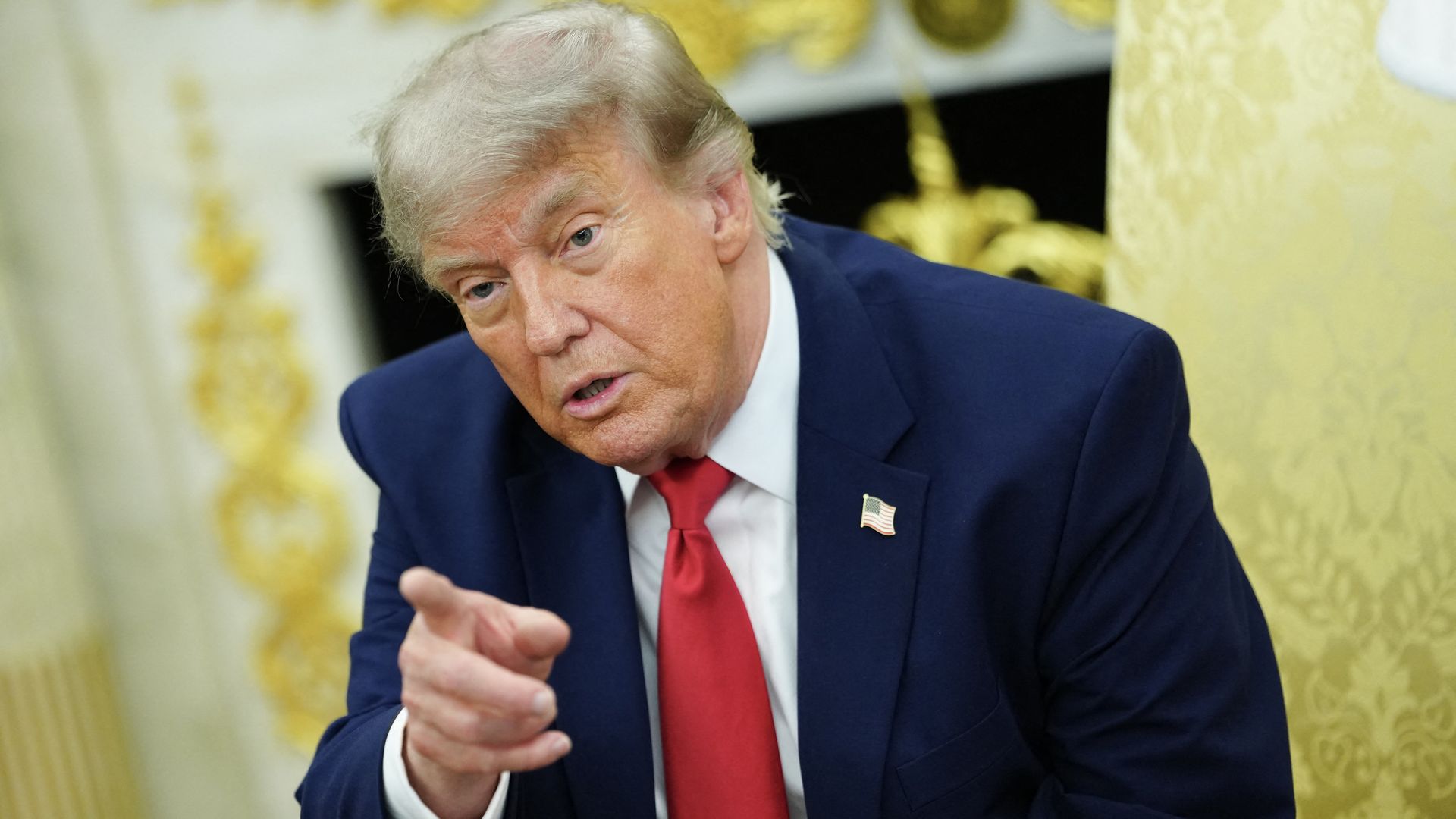 Close-up of President Donald Trump in a dark blue suit and red tie, pointing with his finger. He wears an American flag lapel pin, with a gold ornate background.