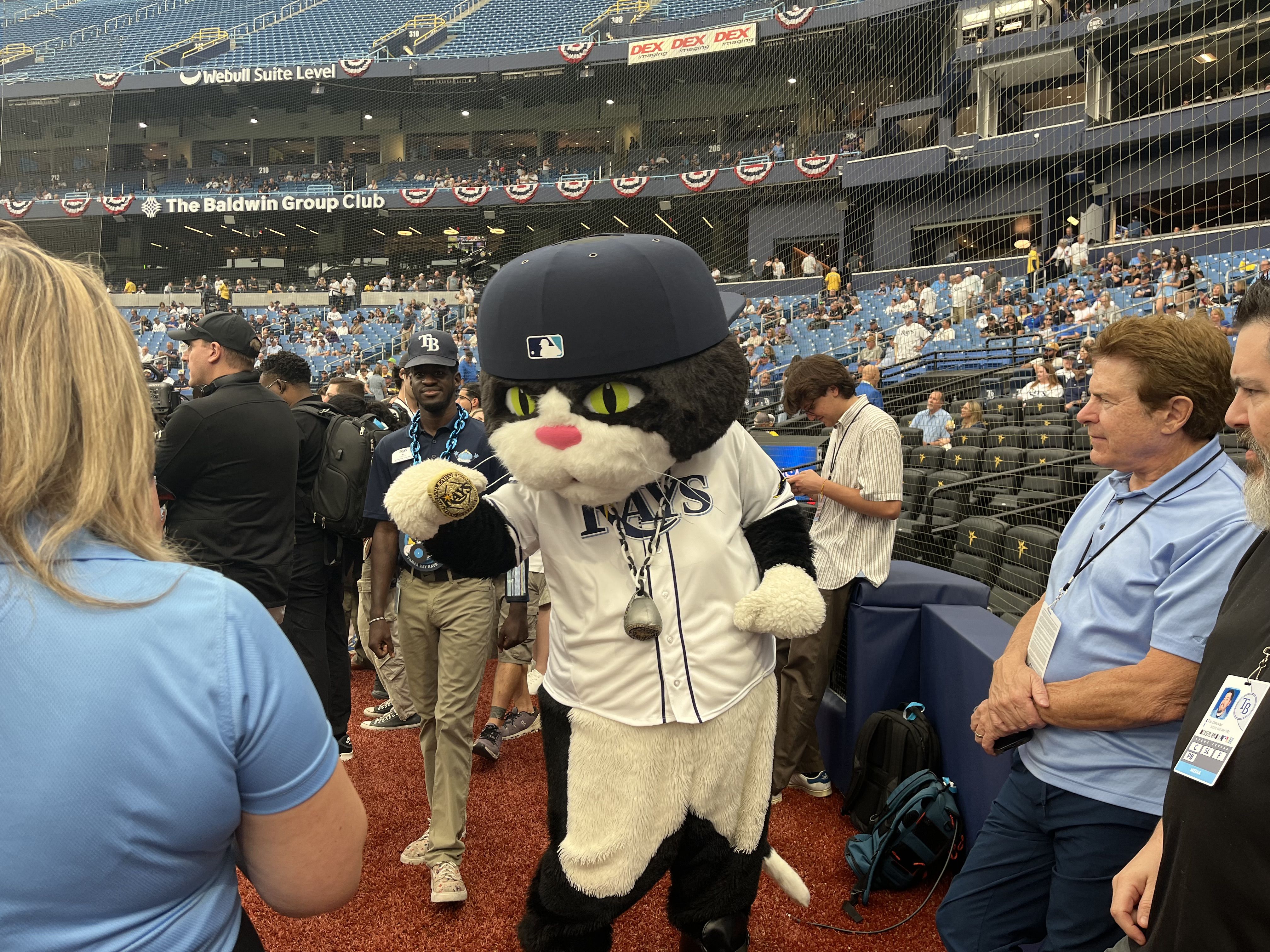 The Tampa Bay Rays mascot in a white jersey and navy helmet poses with fans near the dugout as red, white, and blue bunting decorates the stands at a baseball stadium.