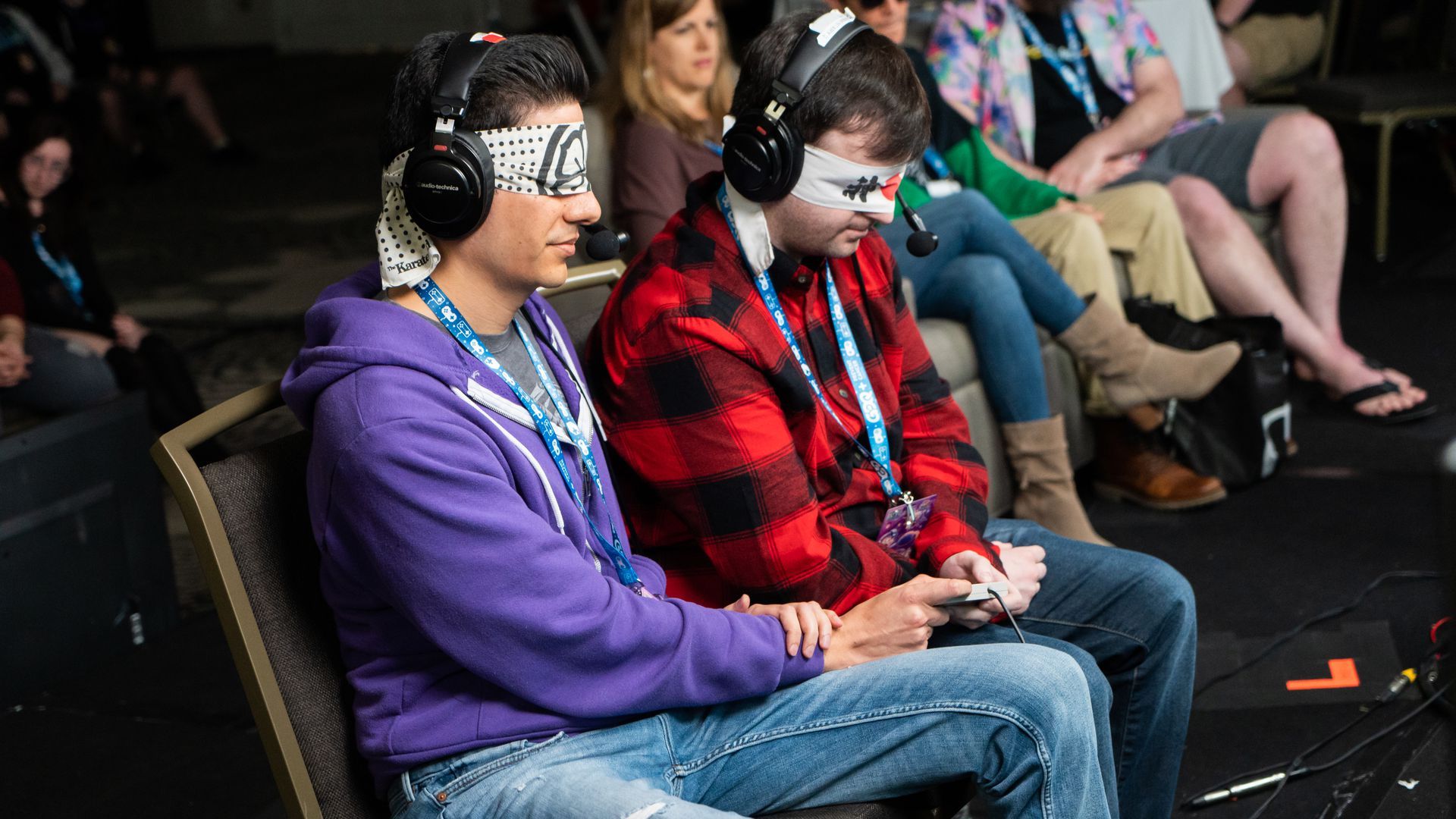 Photo of two blindfolded gamers wearing headphones and holding small rectangular consoles