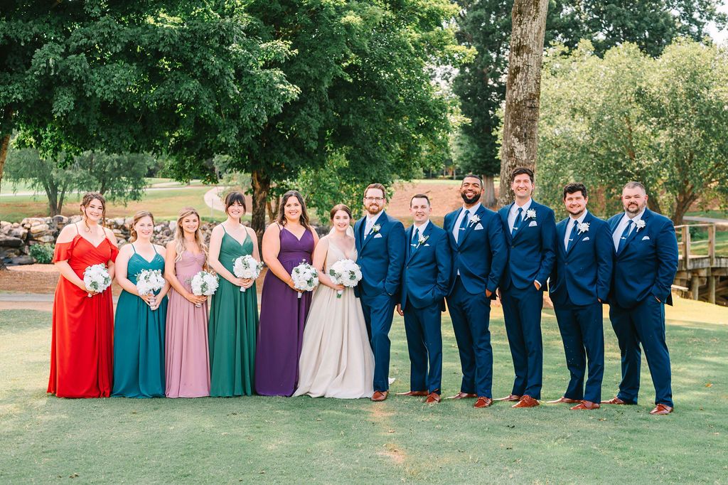 Wedding party outdoors with six bridesmaids in different colored dresses and six groomsmen in blue suits, standing on grass with trees and a bridge in the background.