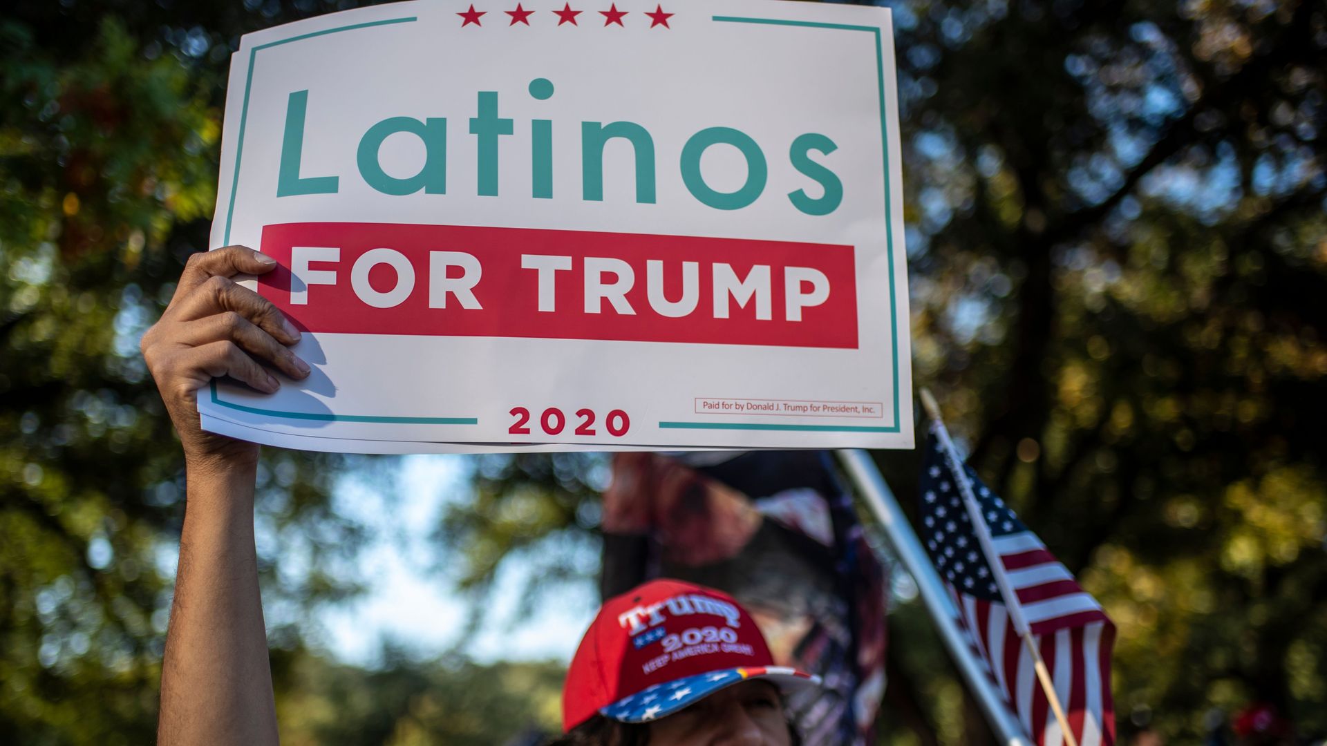 Photo of a person wearing a red Trump 2020 cap and holding up a "Latinos for Trump" sign at an outdoor protest