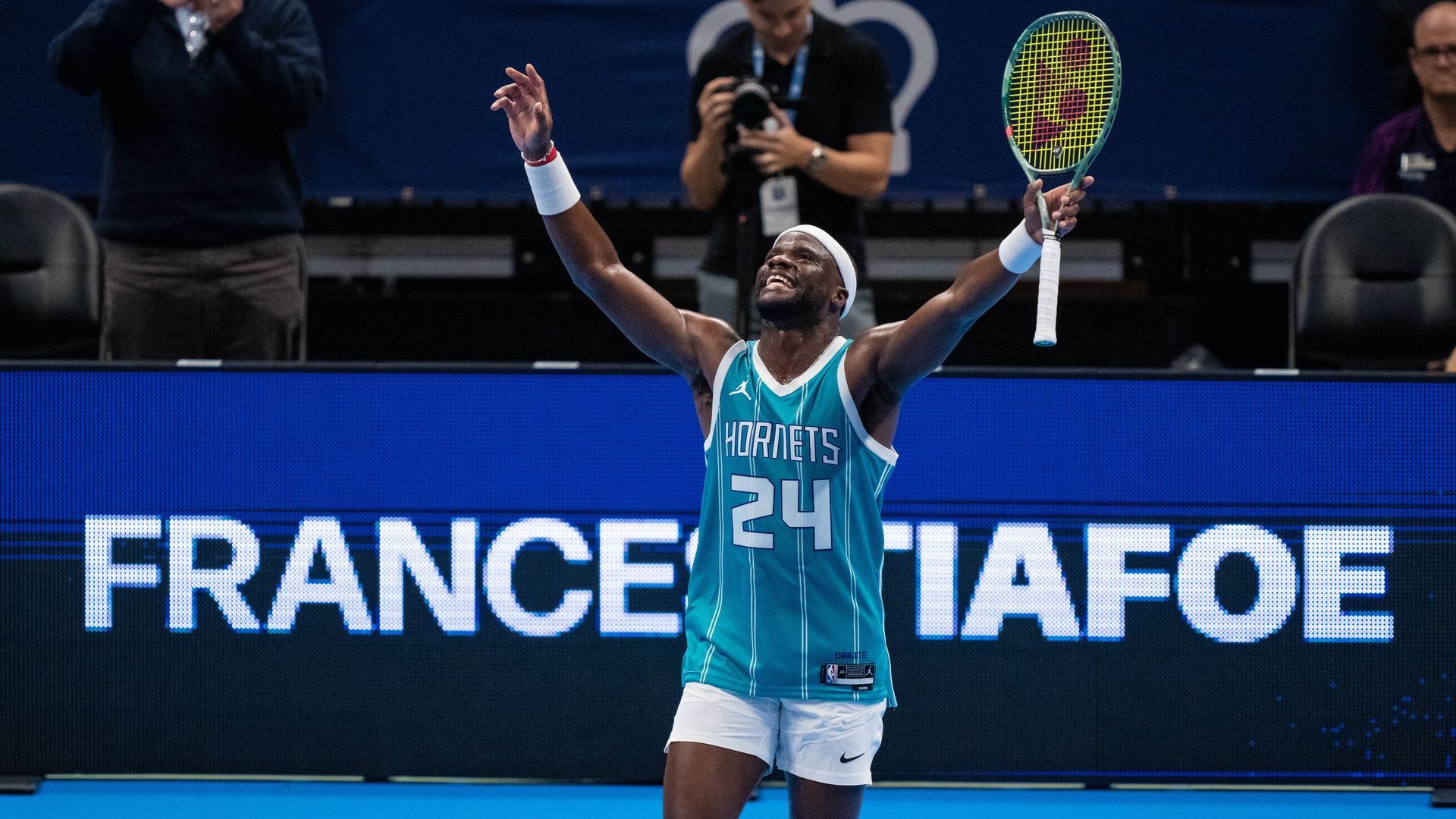 Frances Tiafoe in teal Hornets basketball jersey and white shorts holding a tennis racket, raising arms and smiling on indoor tennis court with blue background.