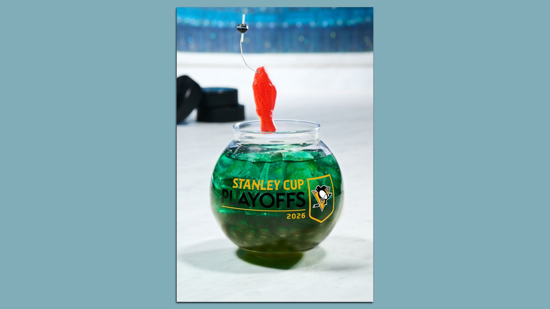 Green drink in a rounded glass on ice, with a red gummy fish on a hook above; the glass shows "STANLEY CUP PLAYOFFS 2026" and a Penguins logo, with a blurred hockey rink behind.