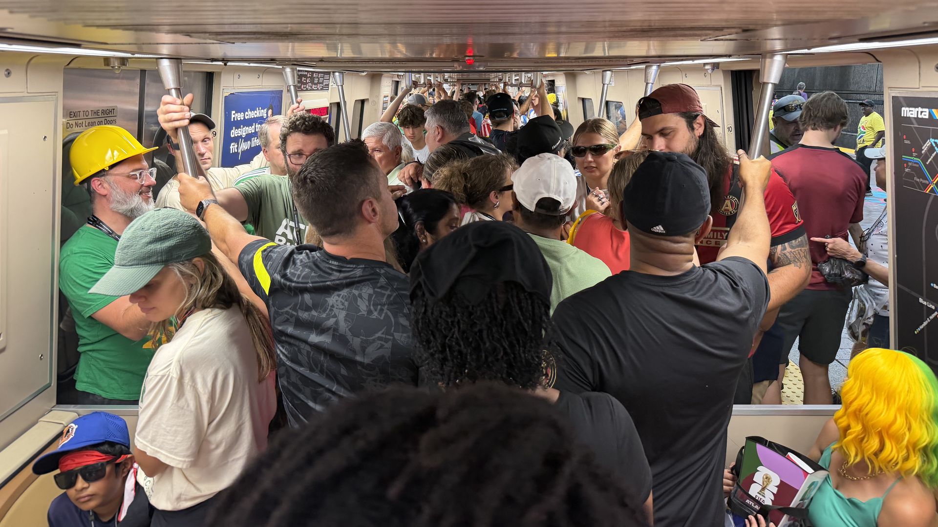 Inside a packed subway car, numerous passengers stand shoulder to shoulder, gripping poles. Some wear hats and sunglasses; a person with rainbow hair is toward the right, and signs line the walls.