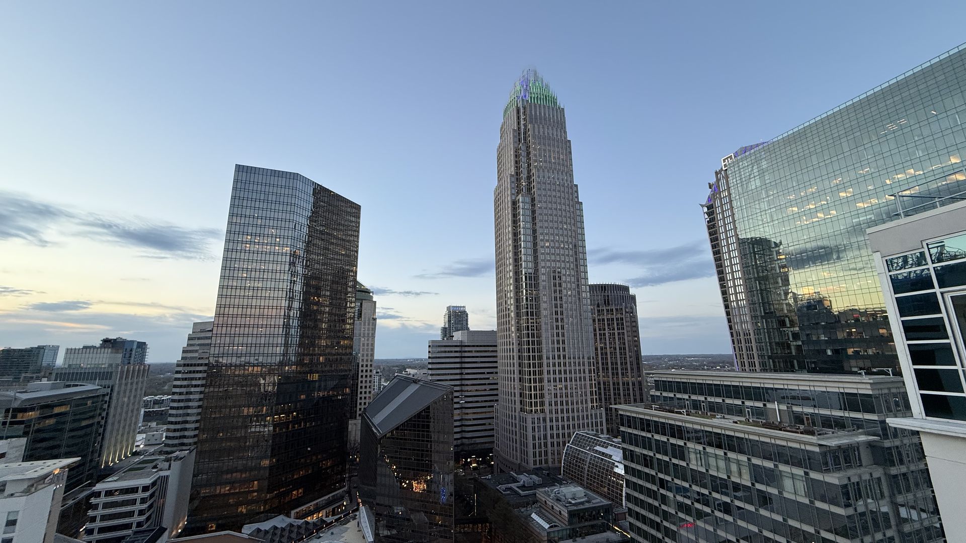 Wide city skyline of glass skyscrapers under a clear blue sky, featuring a central tiered tower with a green-lit crown and reflective surrounding buildings.