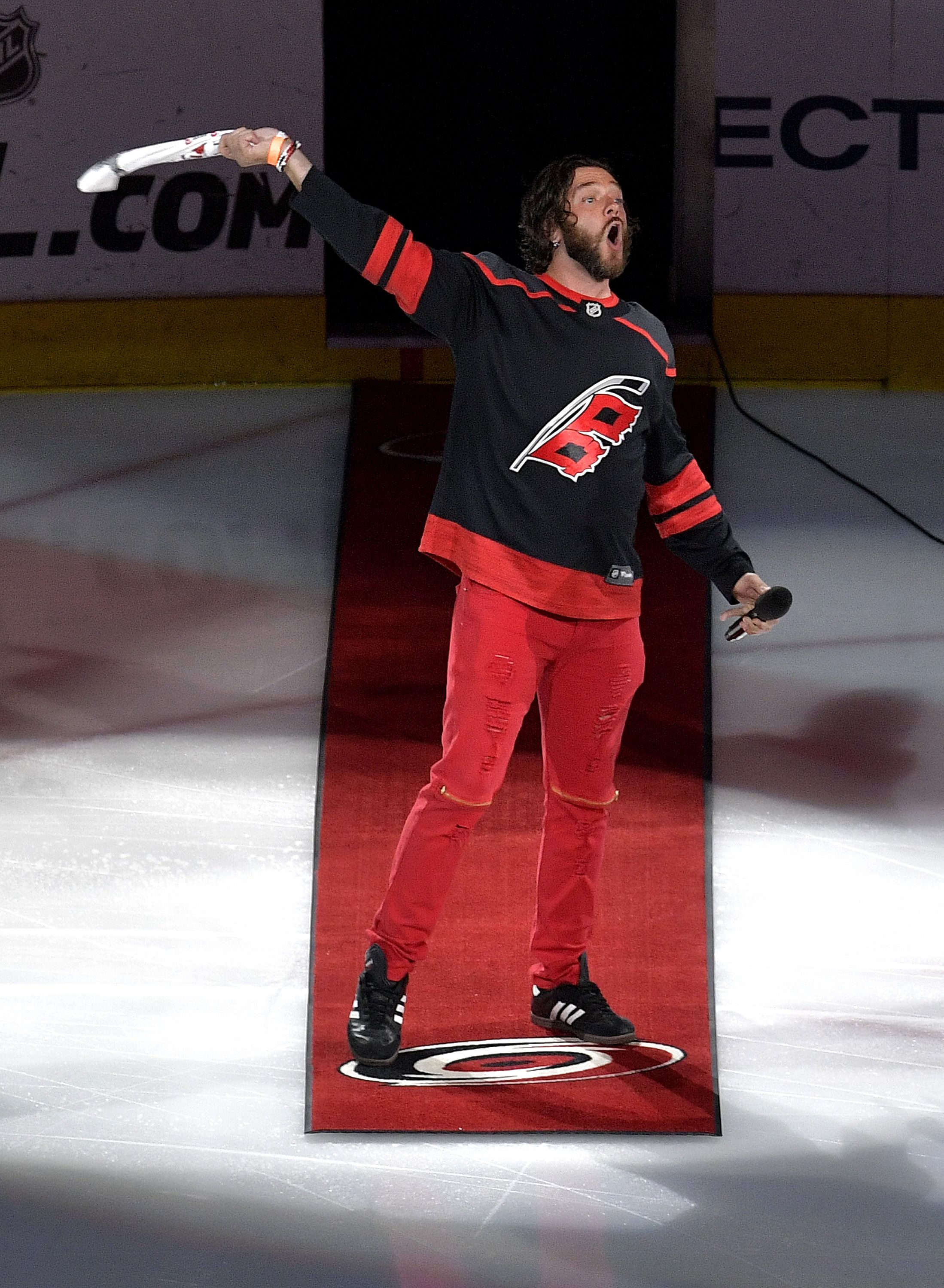 Singer Adam Lee Decker performs prior to an Eastern Conference Finals game in Raleigh during the 2019 NHL Stanley Cup Playoffs. He wears red and black Carolina Hurricanes gear on the ice, waving a towel and holding a microphone.