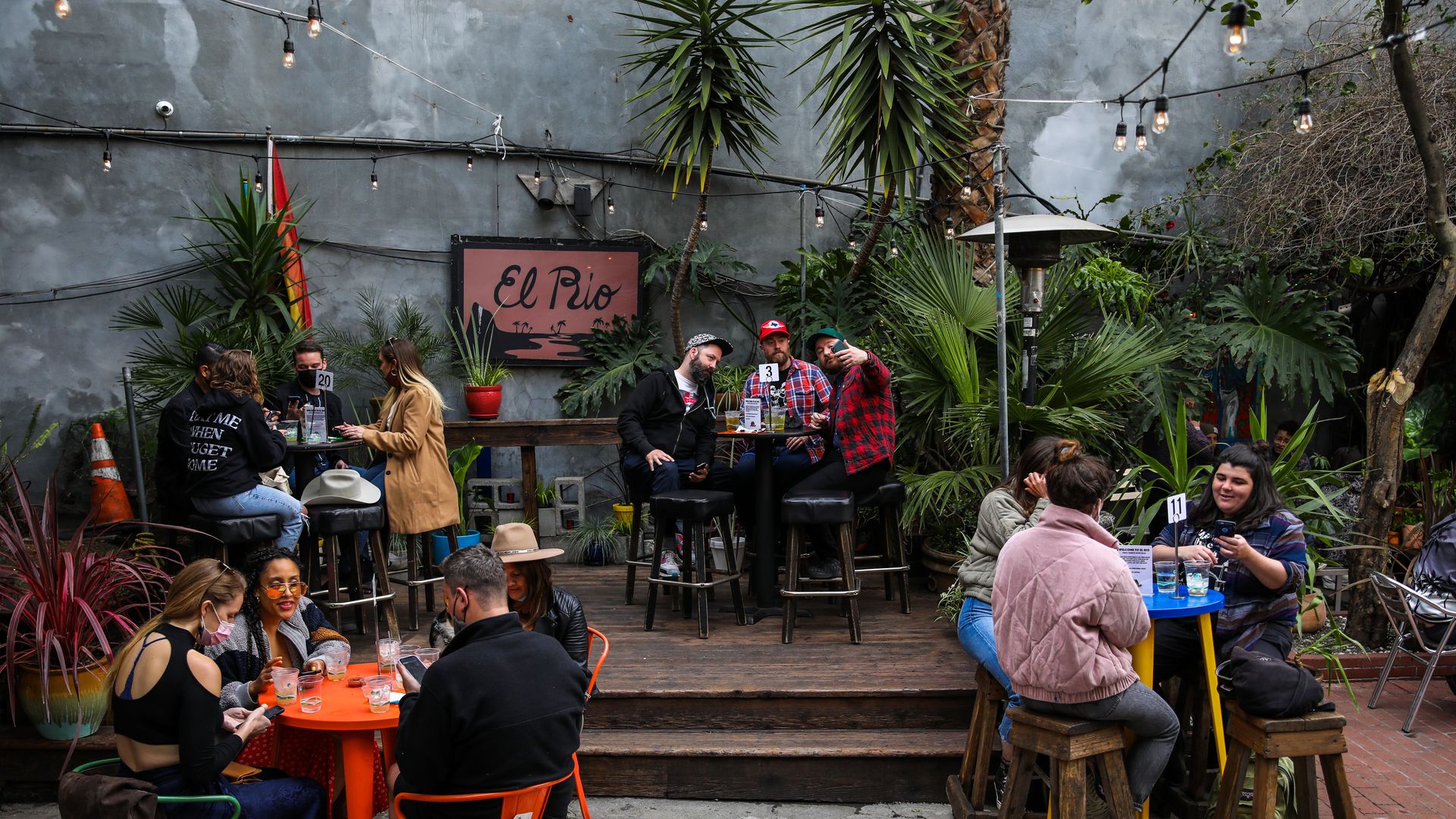 Outdoor cafe with string lights and lush plants along a gray wall. People sit at wooden tables; a pink sign reading "El Rio" is visible in the back.