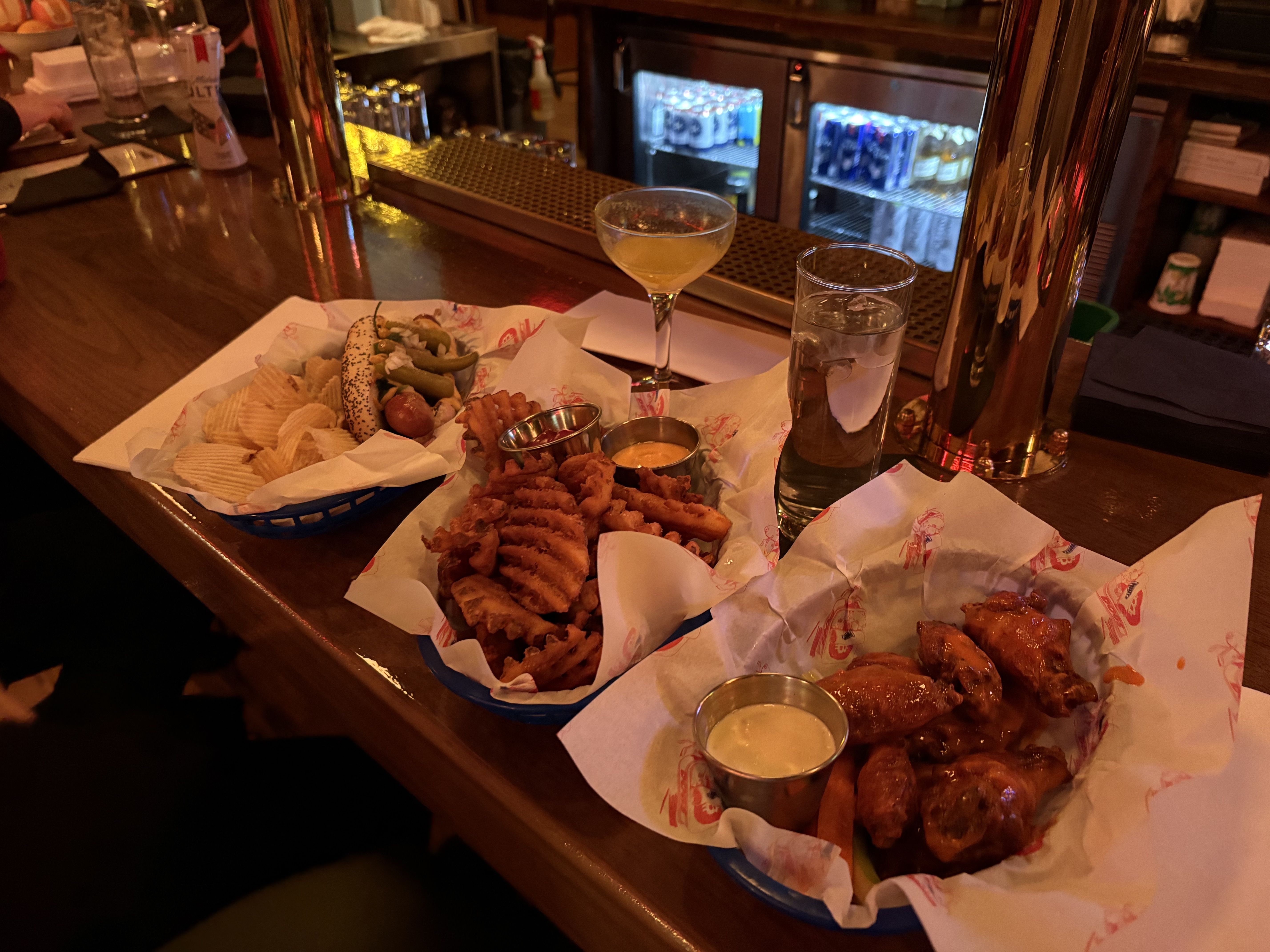 Bar counter with three blue baskets holding potato chips with a hot dog, waffle fries with sauces, and sauced chicken wings with dipping sauce, next to drinks in dim lighting.